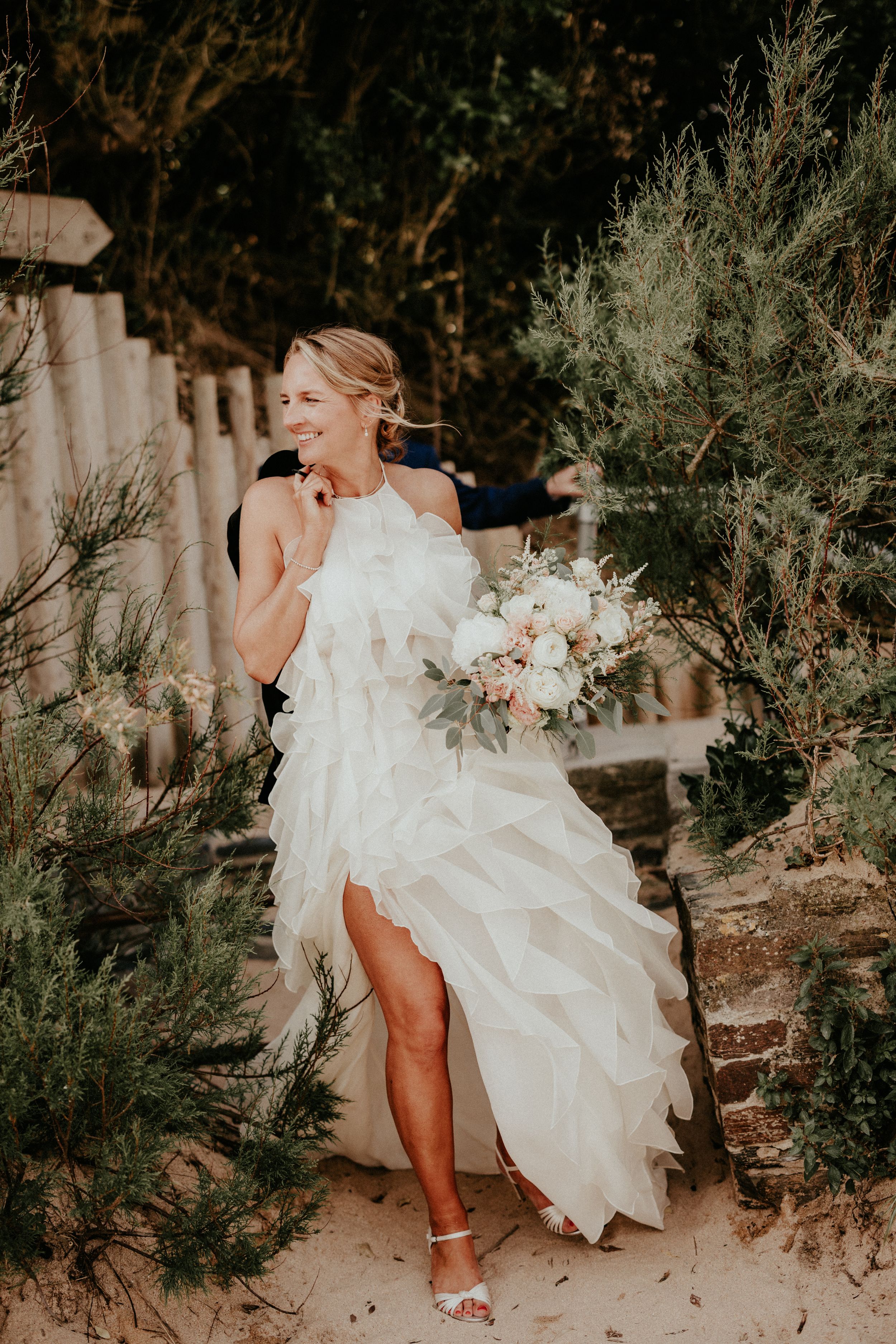 Smiling bride in ruffled white wedding gown holds floral bouquet among greenery on sandy path.