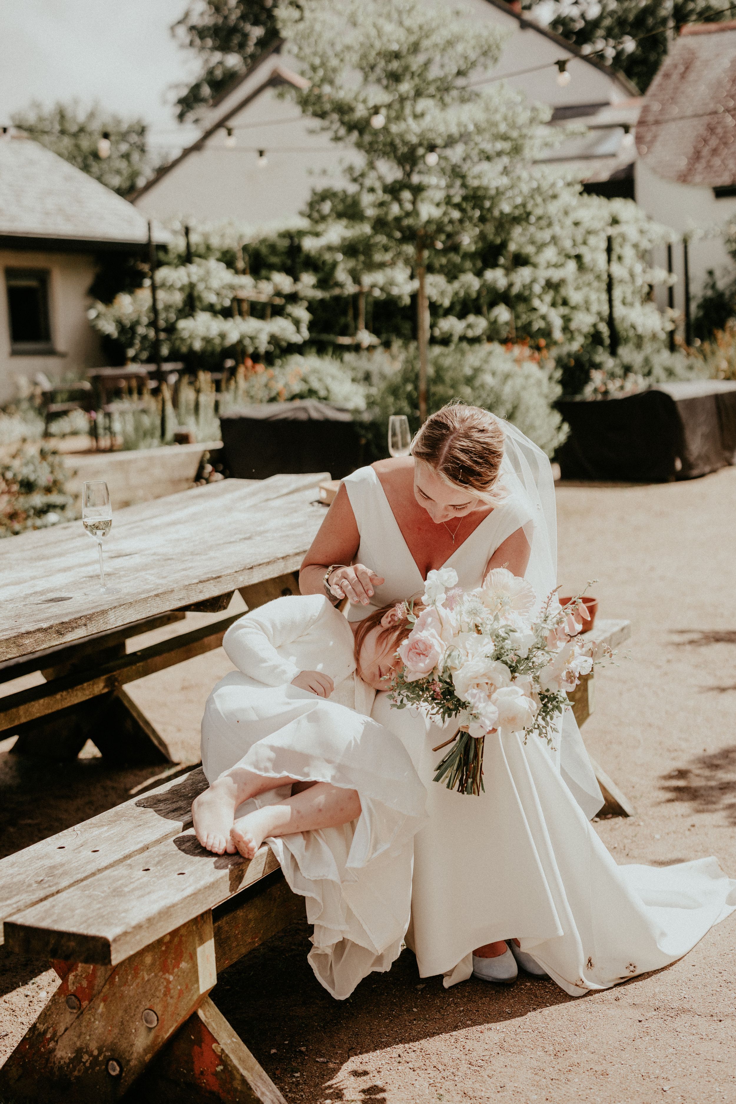 Bride in white gown holding pink bouquet sits on wooden bench in garden wedding venue.