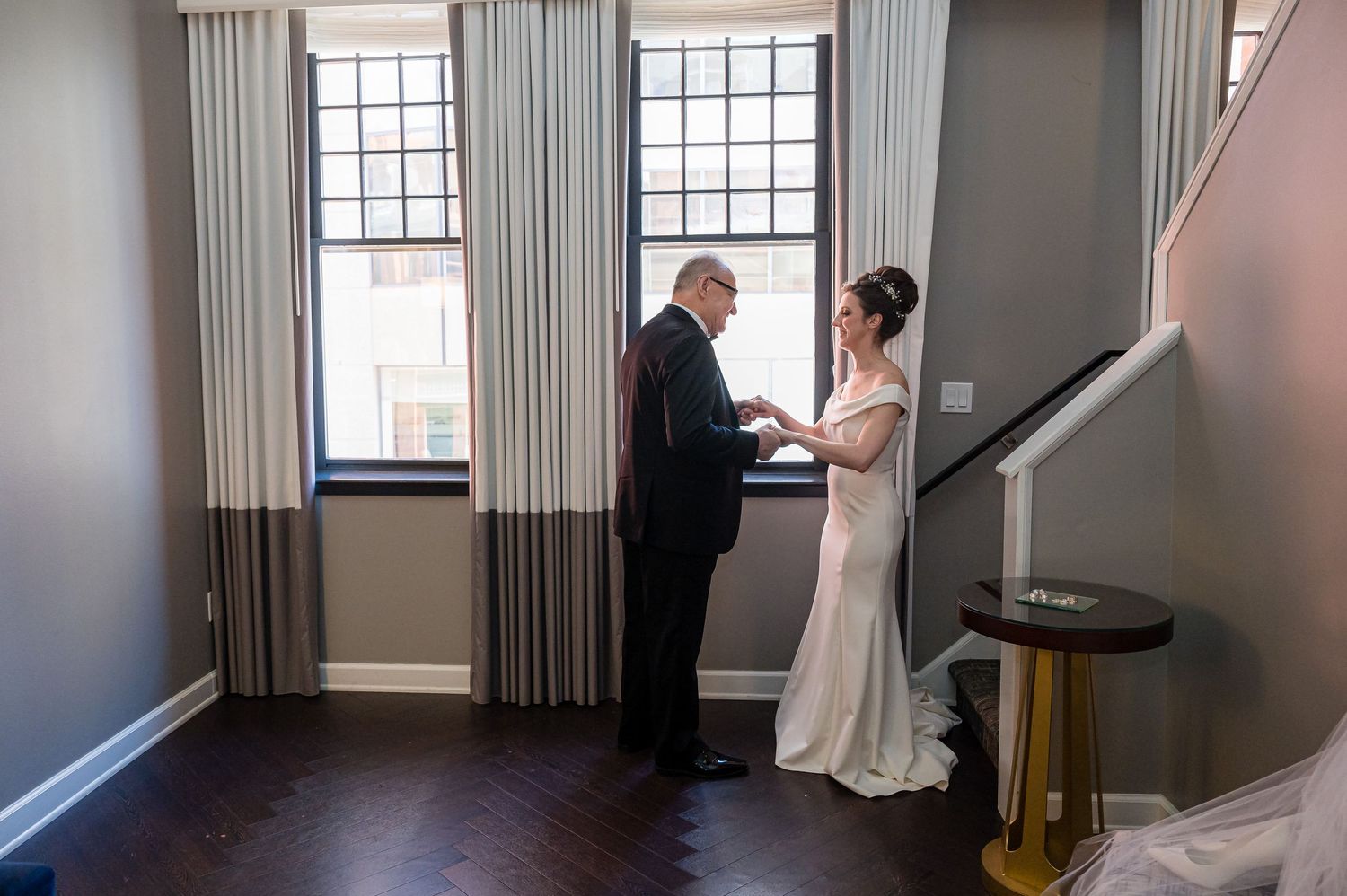 A couple dressed in formal wedding attire share an intimate moment in front of tall windows in an elegant room.