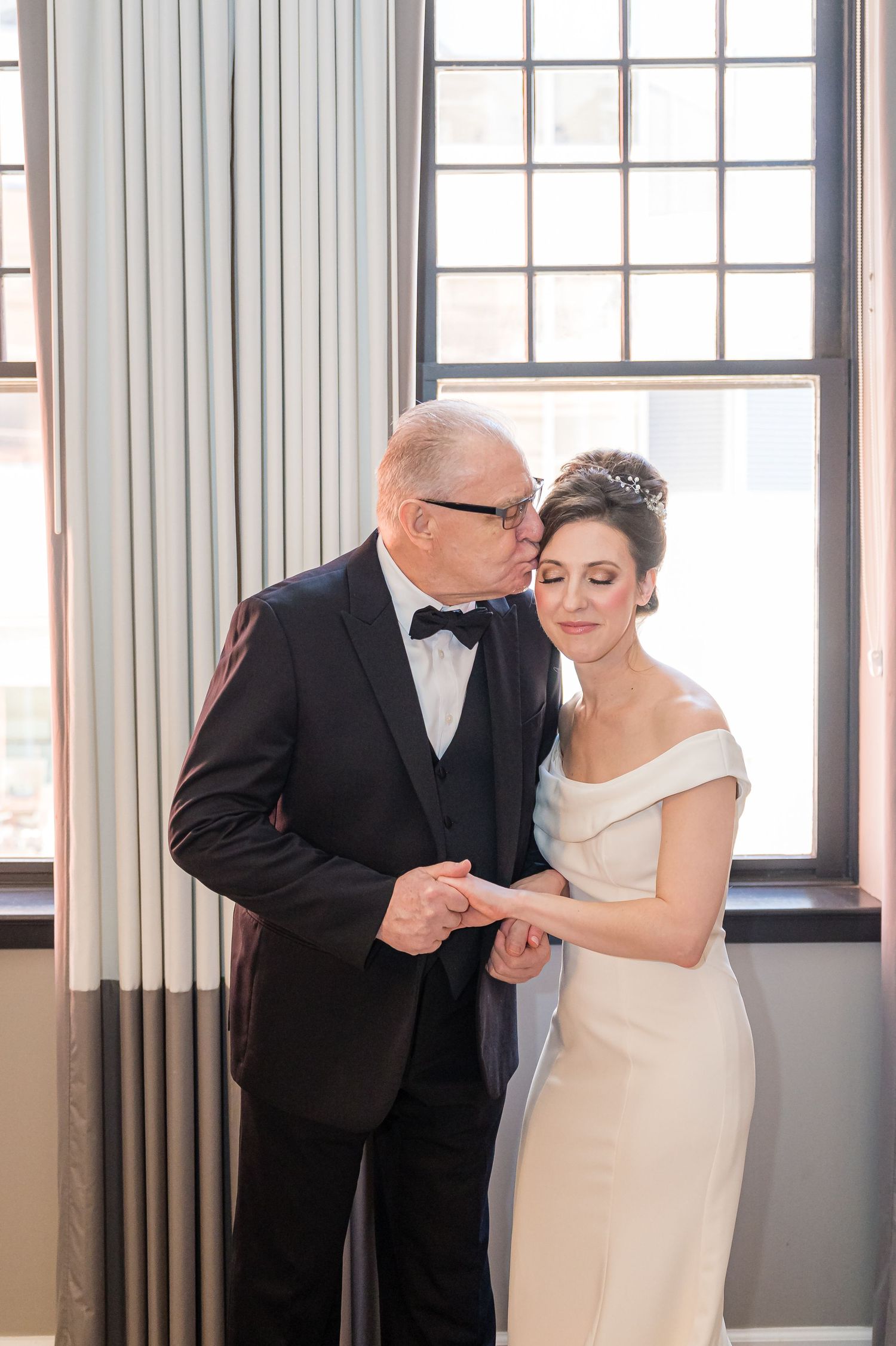 A tender moment shared between a formally dressed pair at an elegant indoor venue with large windows.