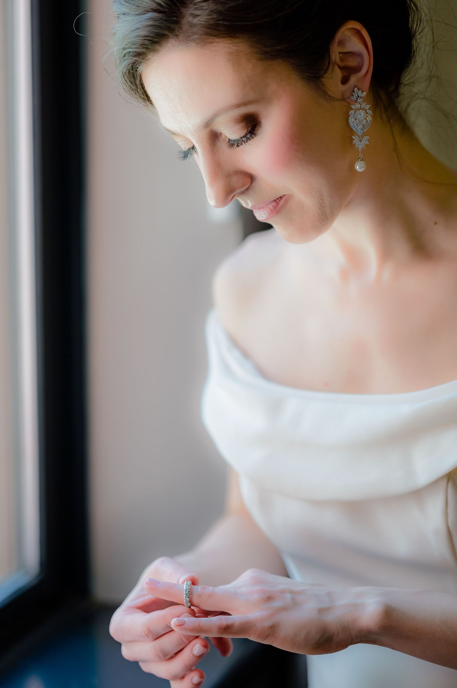A bride in an elegant white gown delicately examines her jewelry near a window with soft natural light.