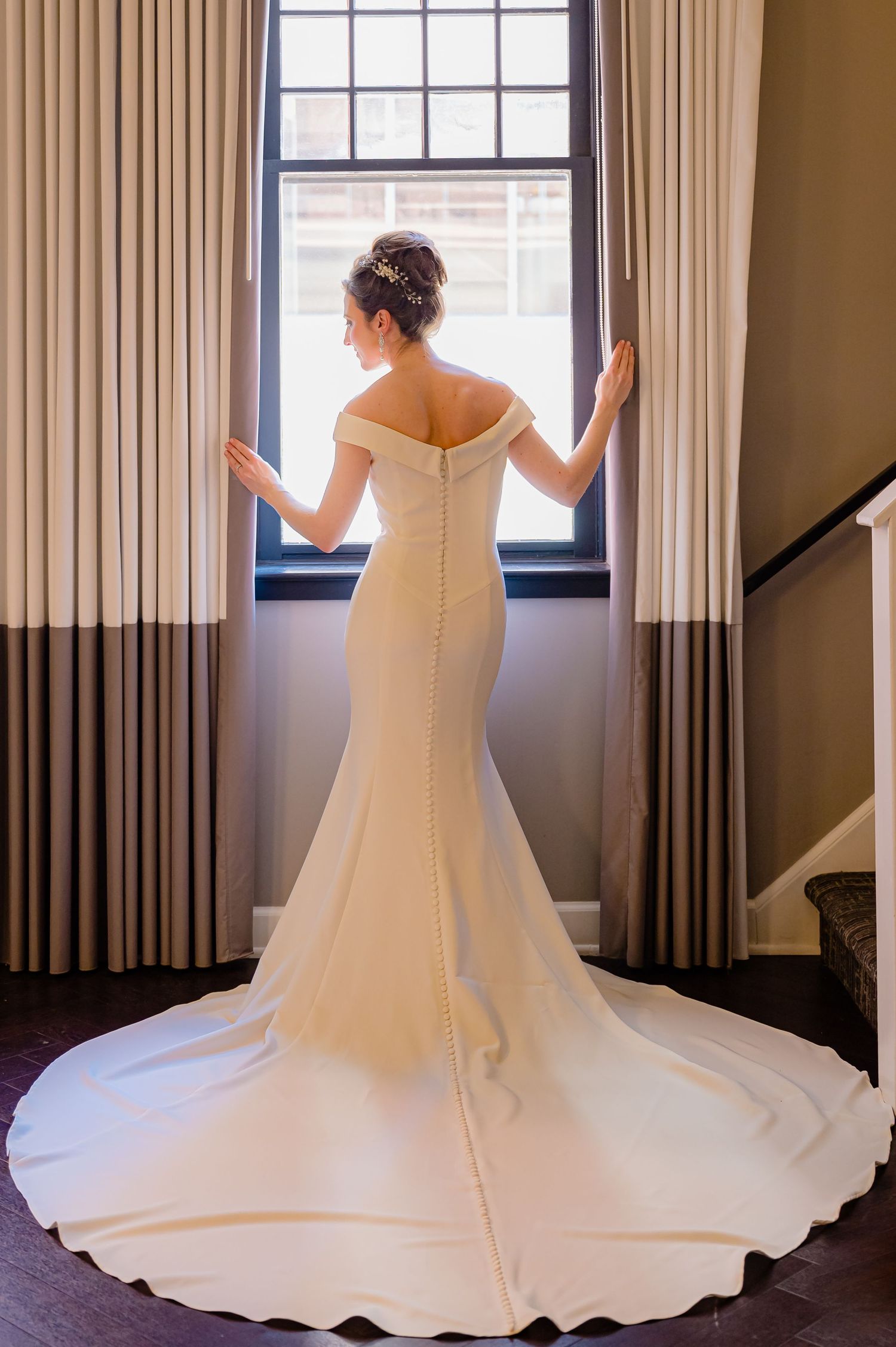A wedding dress with an off-shoulder neckline and flowing train is photographed in different angles against white curtains.