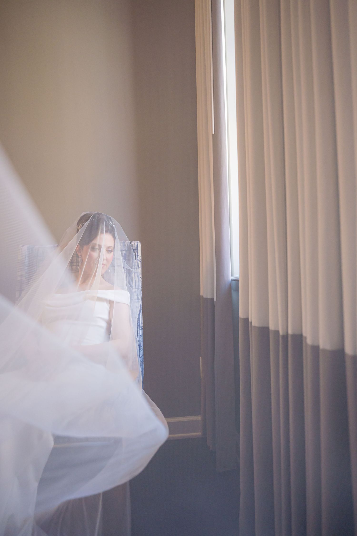Ethereal wedding scene showing flowing white veil and dress near window with soft natural light filtering through curtains.
