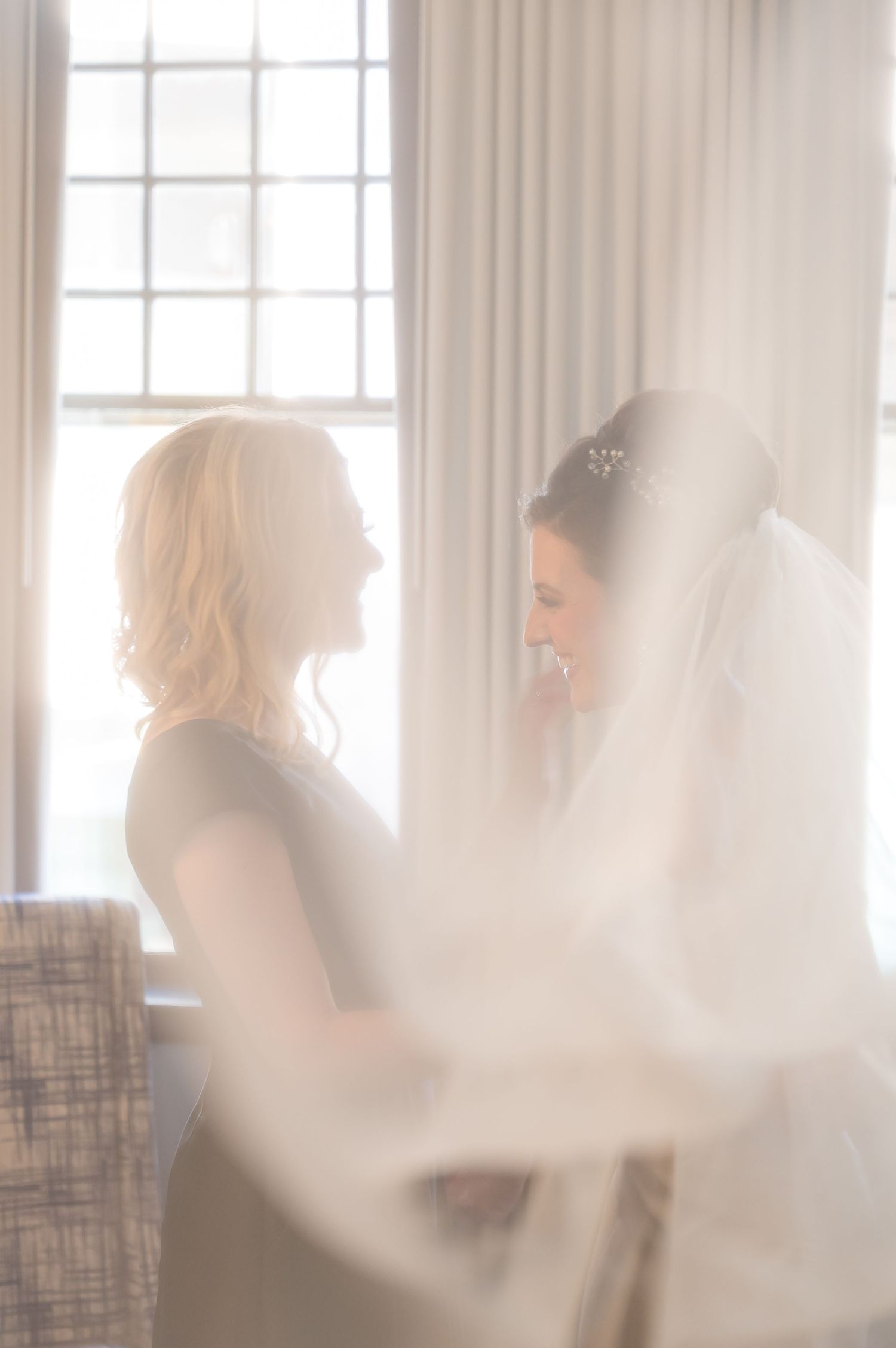 A soft, ethereal bridal scene shows two silhouettes by a bright window with flowing white veil and dreamy lighting.