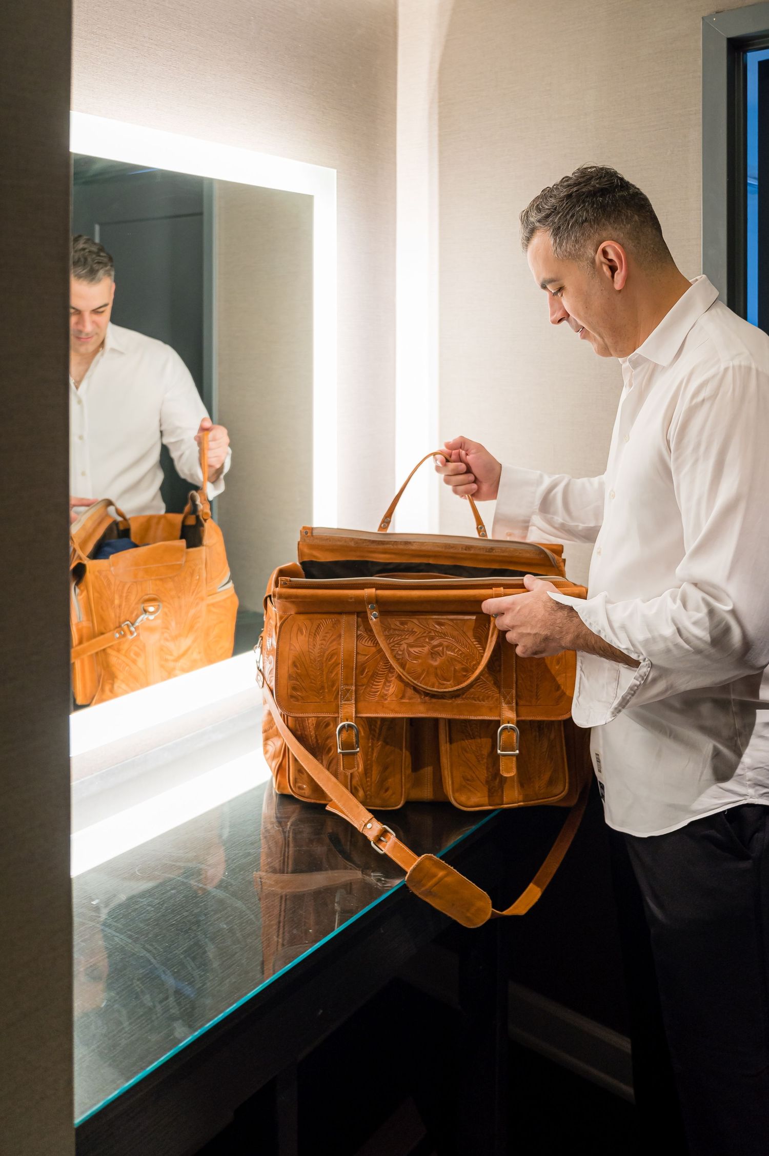 A person in a white shirt examining a brown leather messenger bag near a reflective surface.