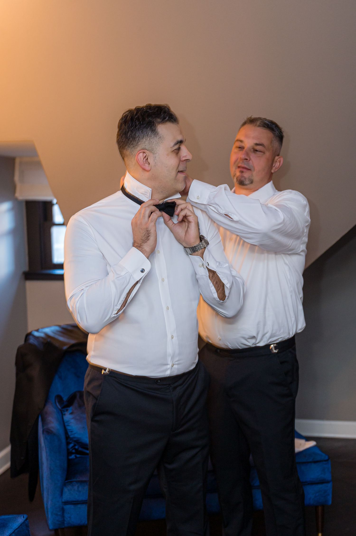 Someone helps adjust a black bow tie on a groom wearing a crisp white dress shirt during wedding preparations.