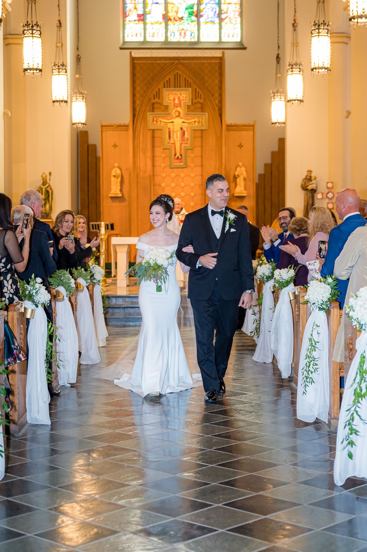 A couple walks down the church aisle during a wedding ceremony with stained glass windows and decorated pews.