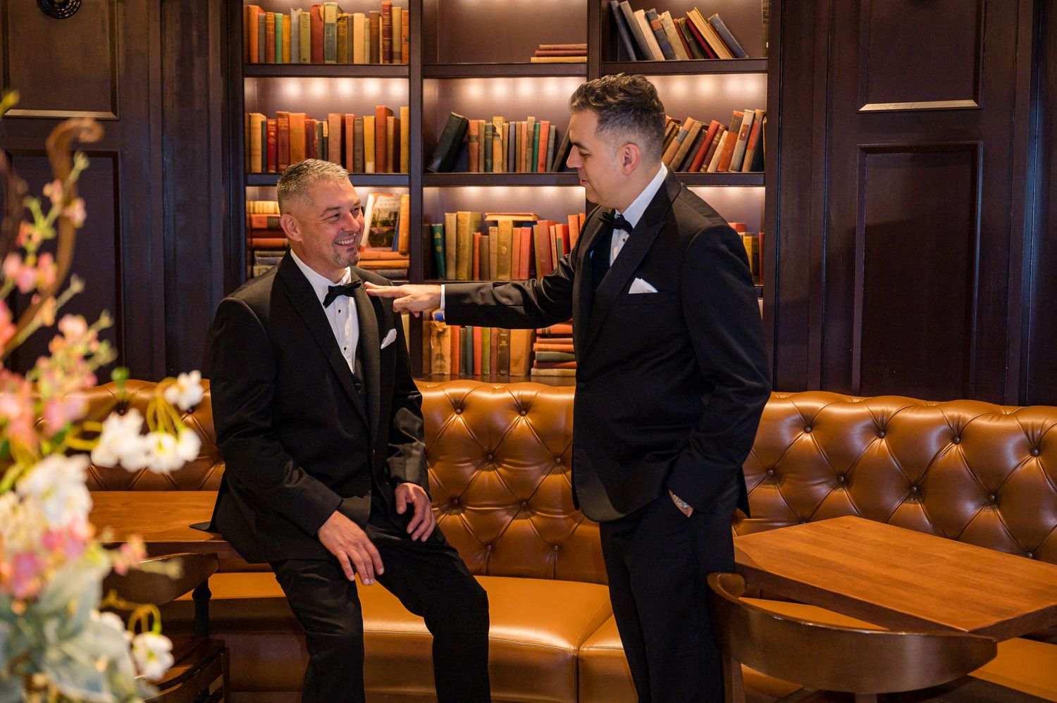 People gather in an elegant library lounge with leather seating and wooden bookshelves glowing in warm lighting.