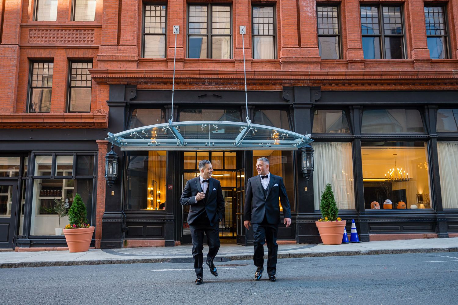Two people walk past a historic red brick building with an elegant glass entrance canopy on a city street.