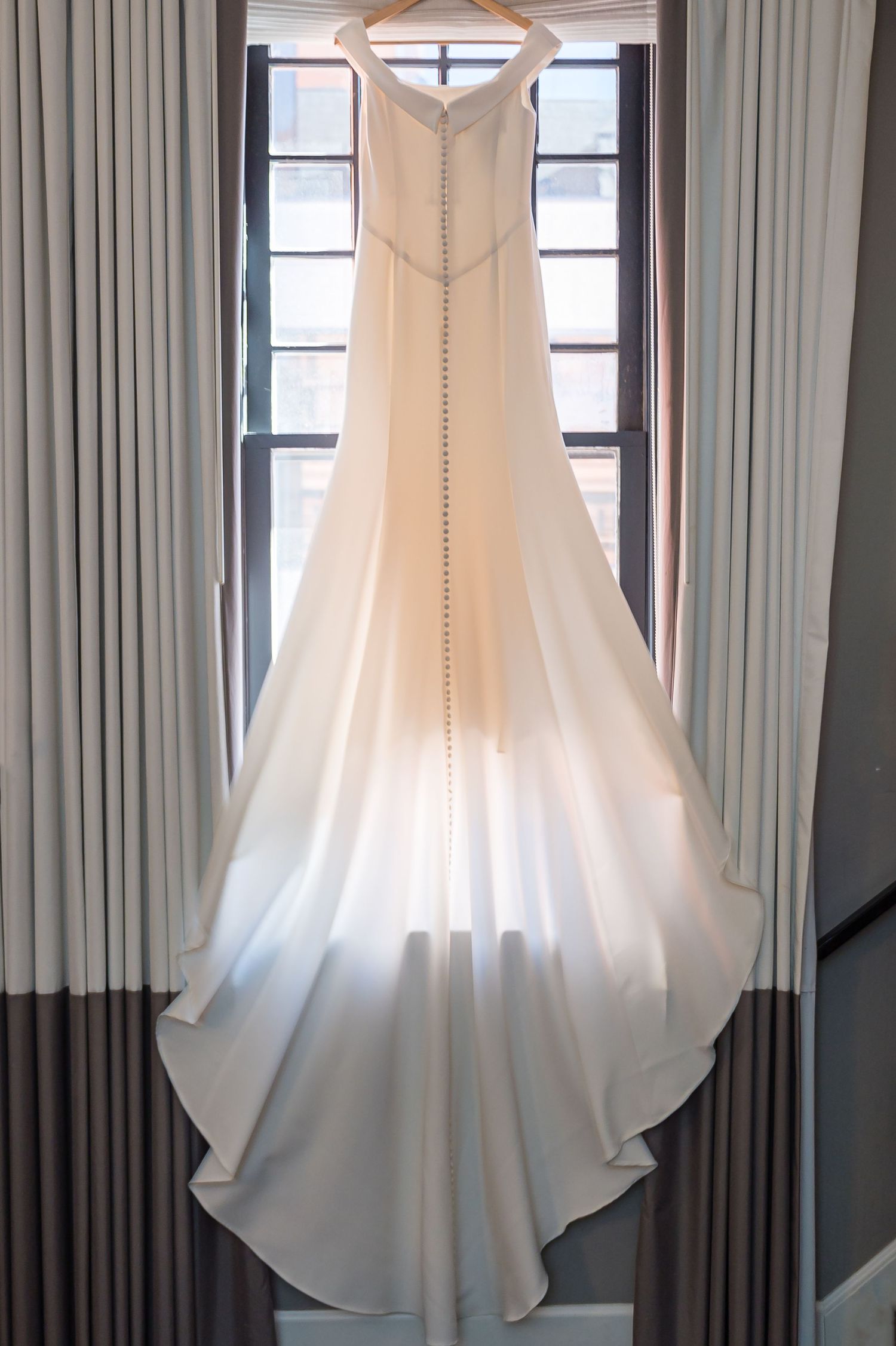 An elegant white wedding dress hanging on display against gray curtains shows its flowing train and detailed silhouette.
