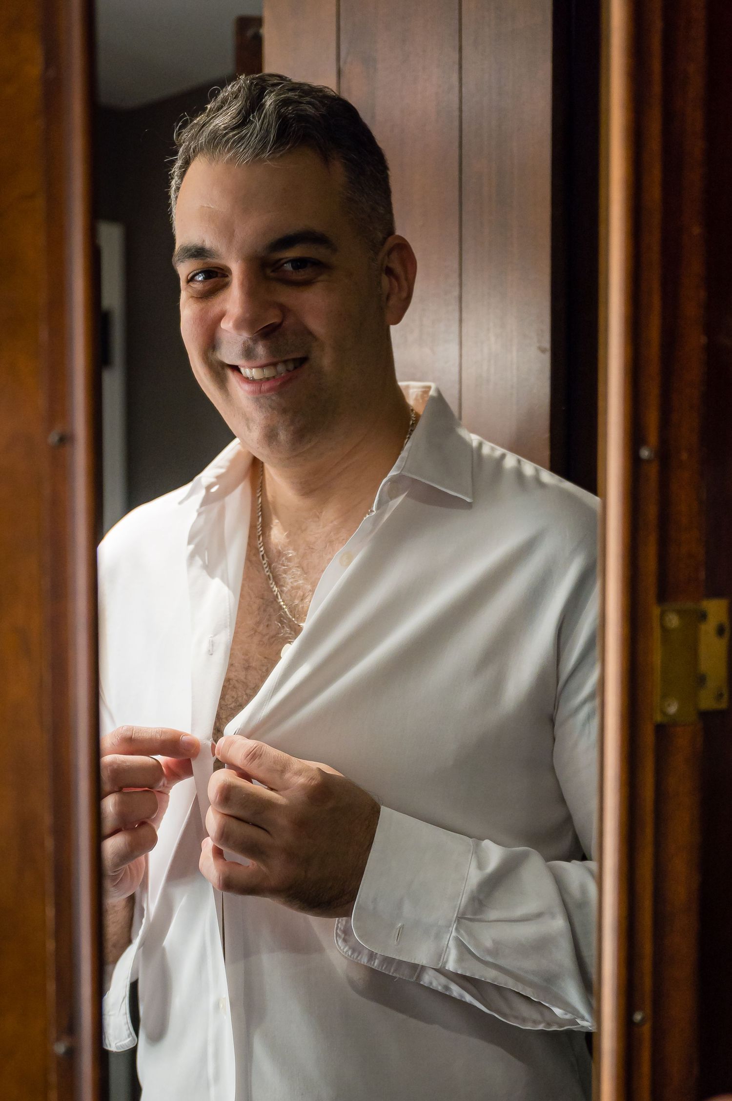 A person in a white button-up shirt smiles while standing in a doorway with wooden trim.