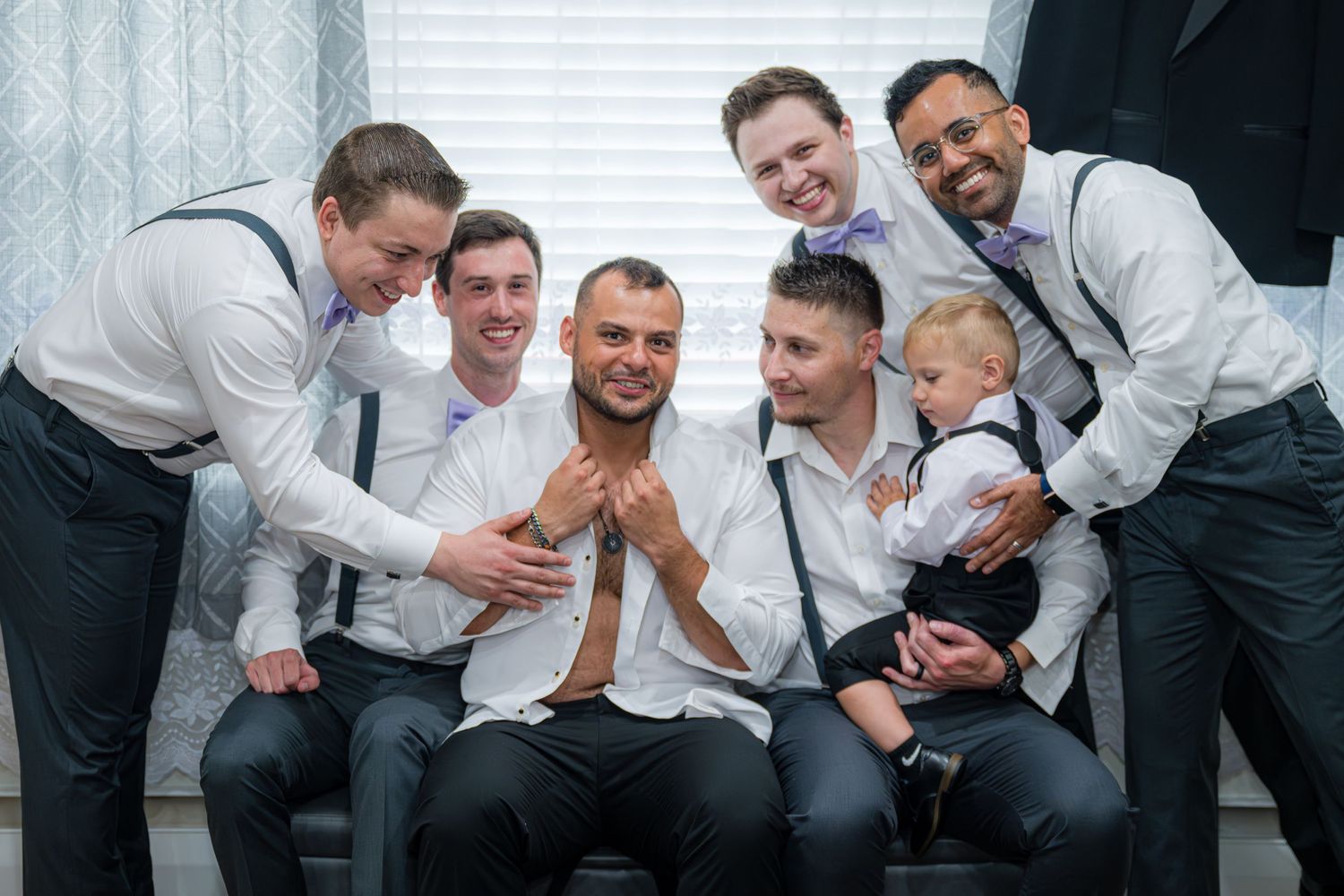 Groomsmen in matching suits share candid laughs while sitting together on a gray couch during wedding preparations.