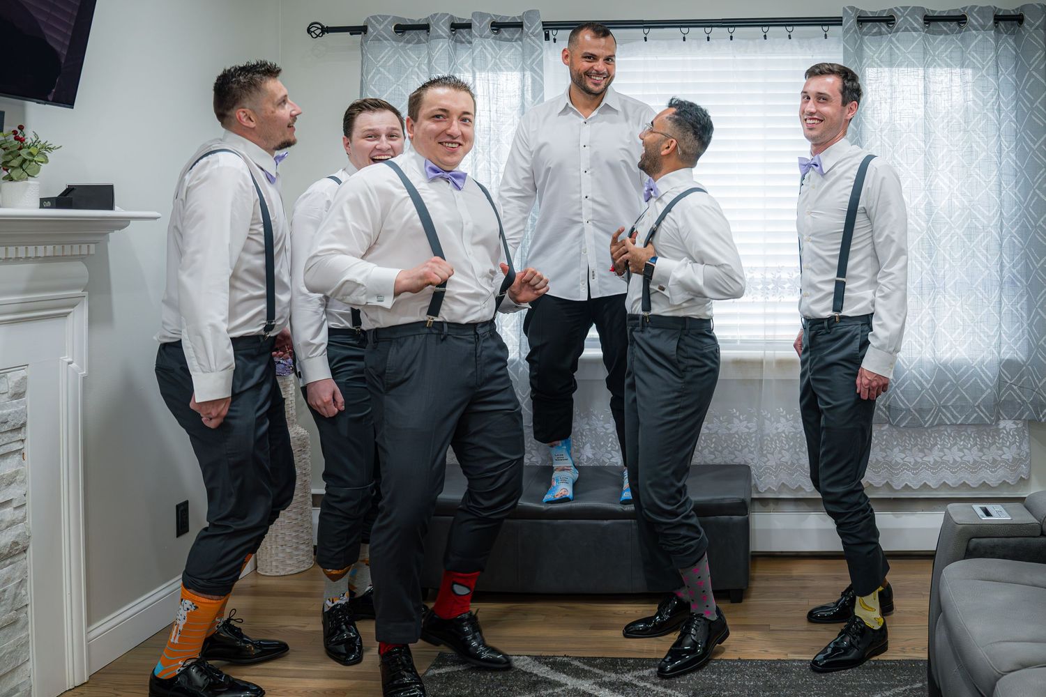 Groomsmen in matching gray and white attire with suspenders and colorful socks pose together in a bright room.