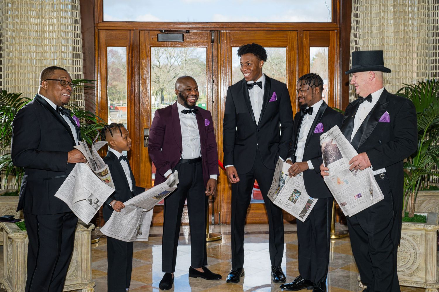 A group of formally dressed people in tuxedos stand together reading newspapers in an elegant room.