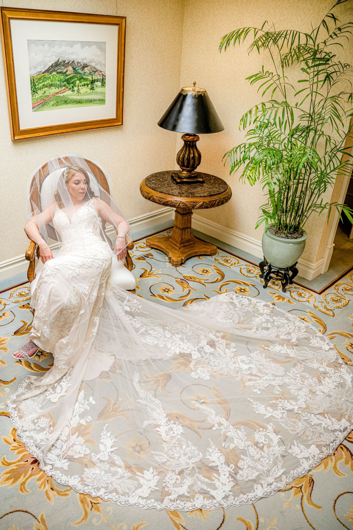 Elegant white lace wedding dress with long train displayed on ornate patterned carpet in luxurious interior setting.
