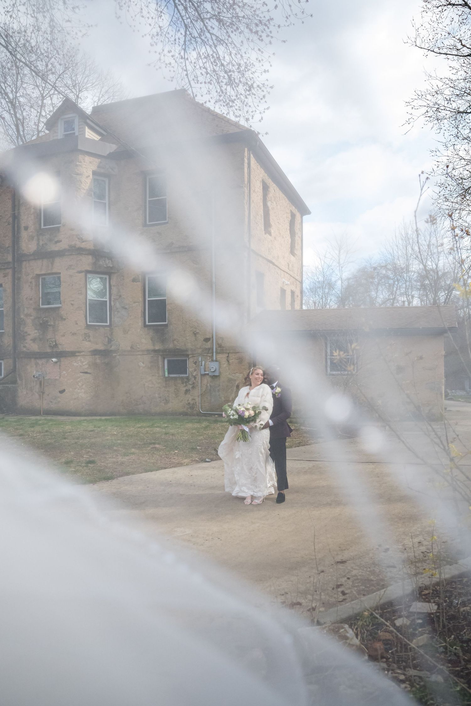 A dreamy winter wedding scene through frosted glass shows a couple in formal attire outside a historic brick building.