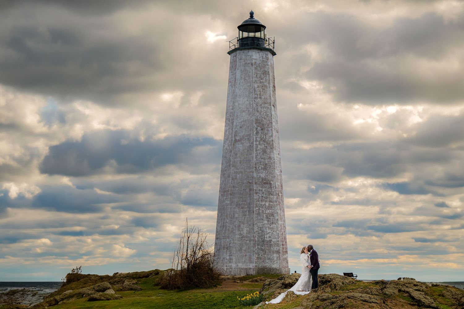 A white lighthouse stands tall against dramatic cloudy skies on a coastal cliff at sunset.