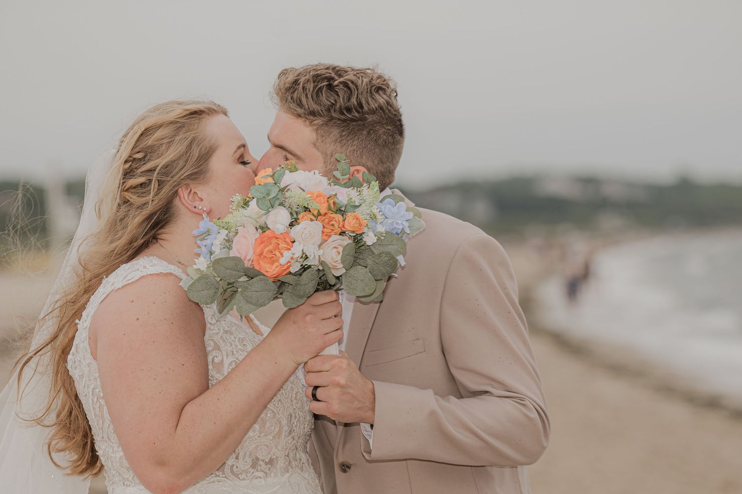Couple in formal attire embracing and kissing on a misty beach with flowers and muted tones.