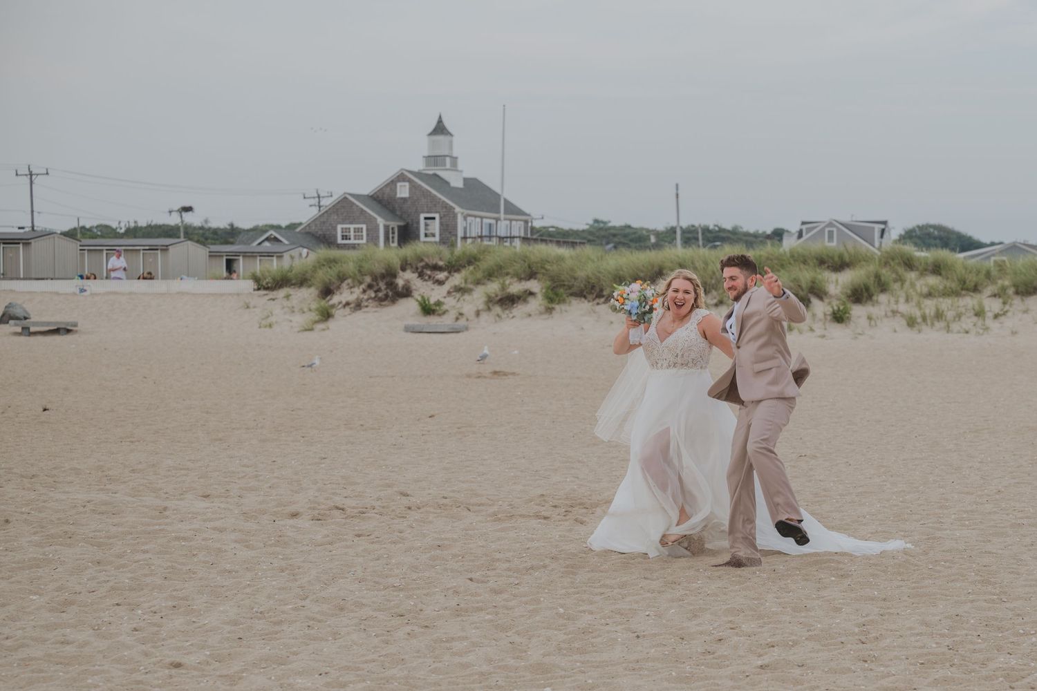 A scenic beach wedding ceremony takes place near a coastal chapel with sand dunes and grasses in the background.
