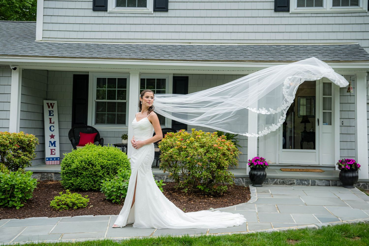 A flowing white wedding dress veil blows gracefully in the wind against a classic New England style home exterior.