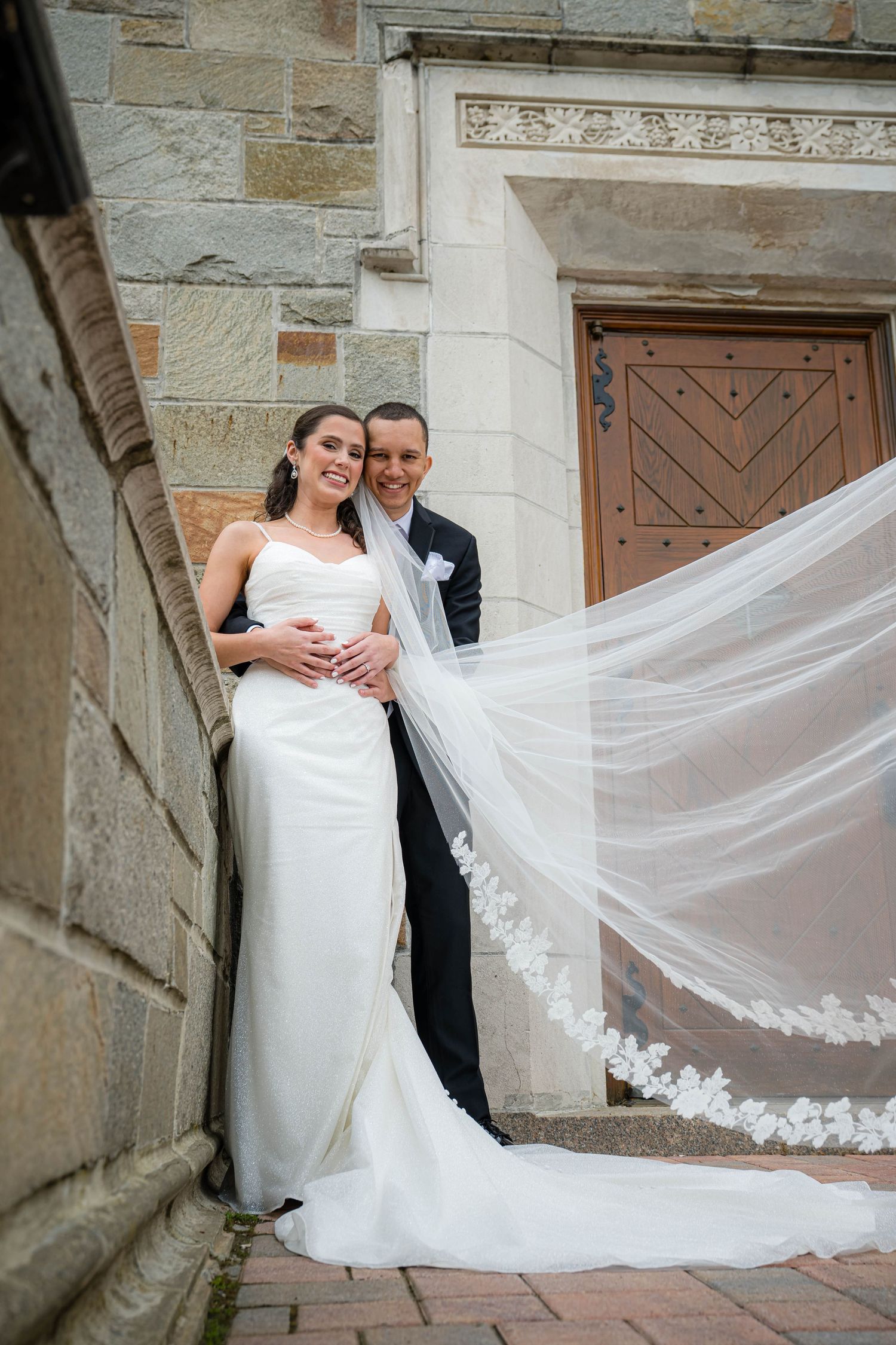 A romantic wedding moment captured on church steps with a flowing lace veil catching the breeze.