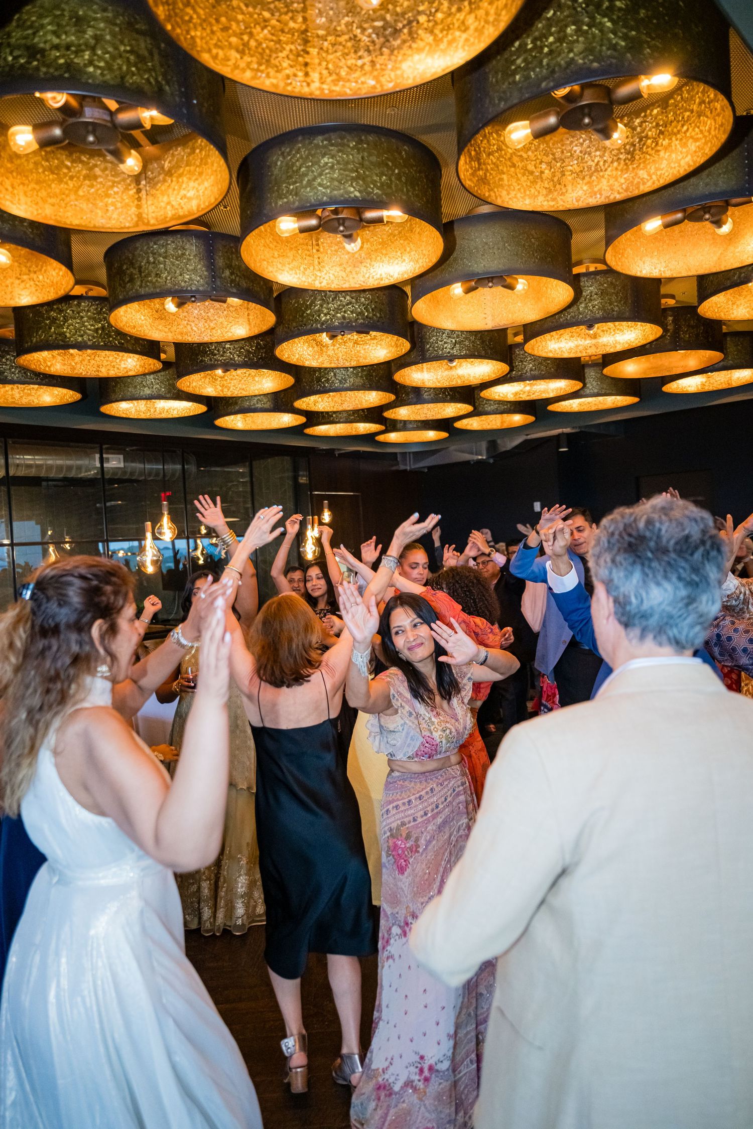Large decorative golden pendant lamps illuminate a dance floor where guests celebrate at an indoor event.