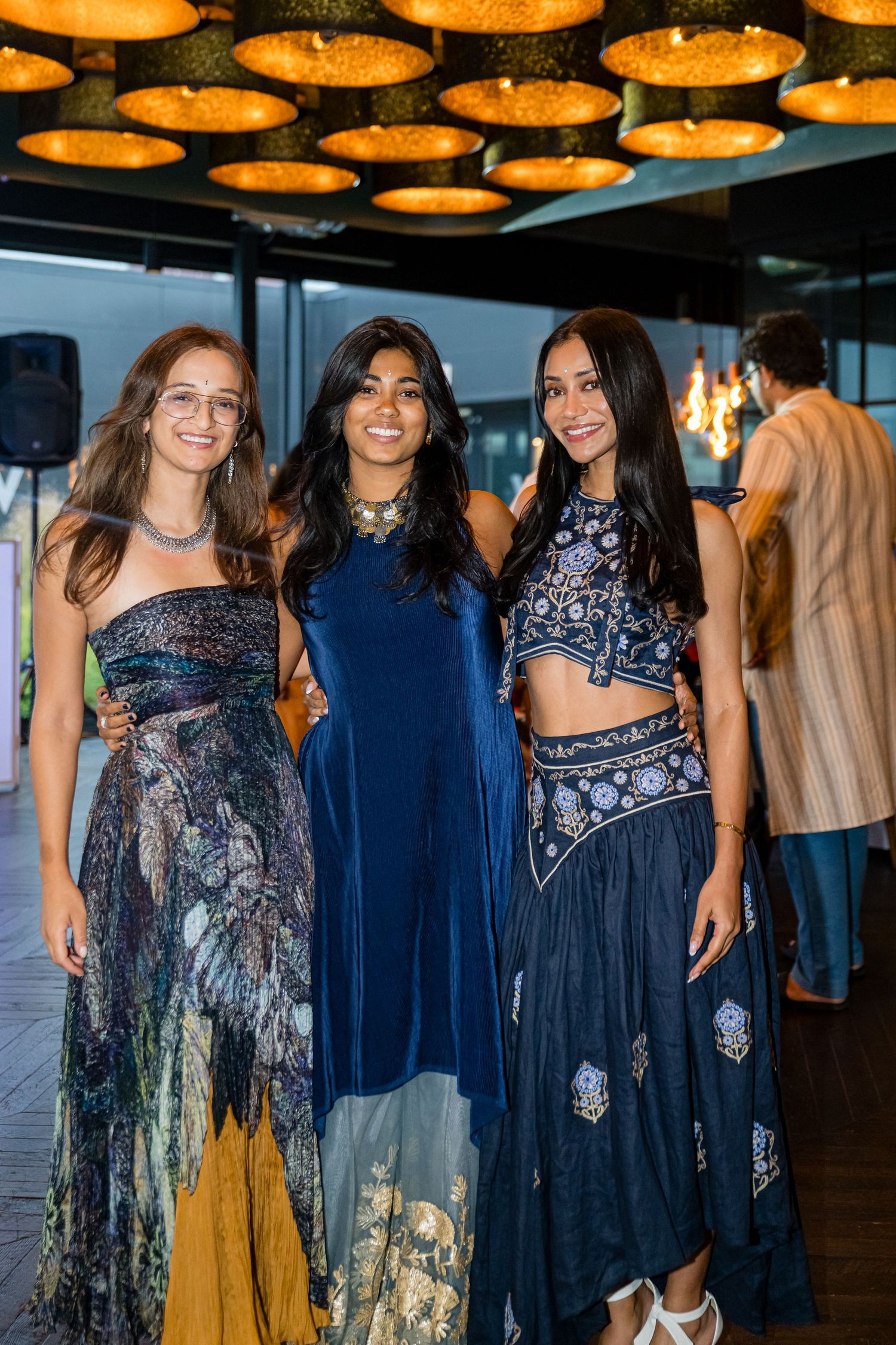 Three women pose at an evening event wearing elegant gowns in blue and navy tones under decorative orange ceiling lights.