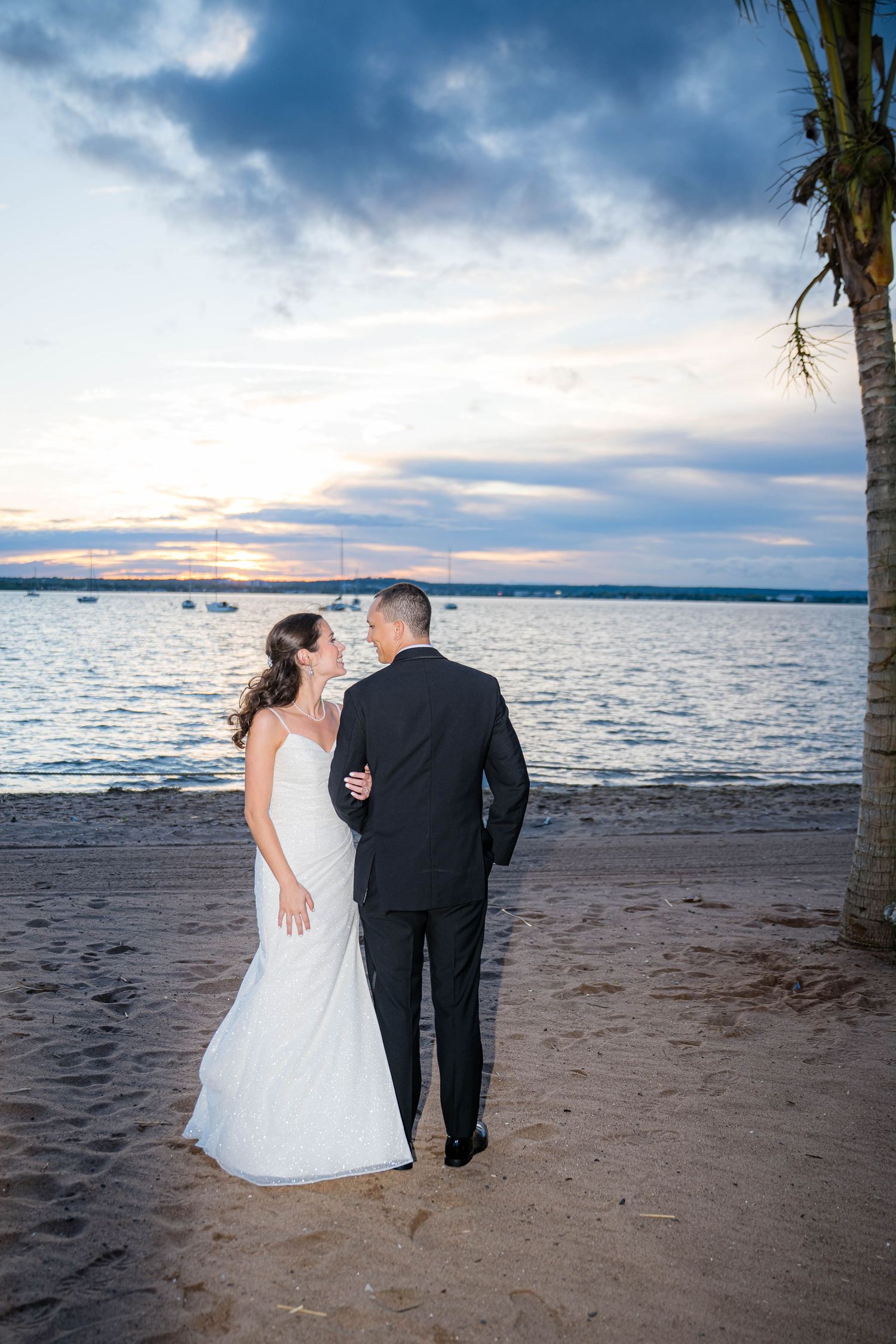 A couple in wedding attire stands on a beach at sunset, with boats and palm trees silhouetted against dramatic clouds.