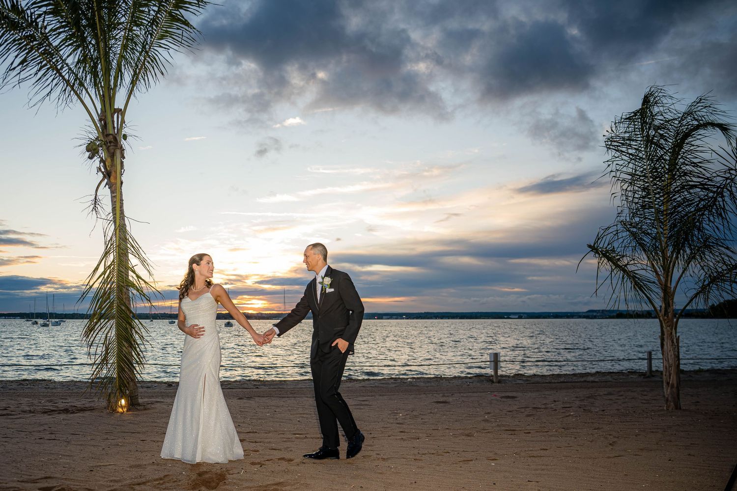 A couple in formal attire dances on a beach at sunset with palm trees and cloudy skies in the background.