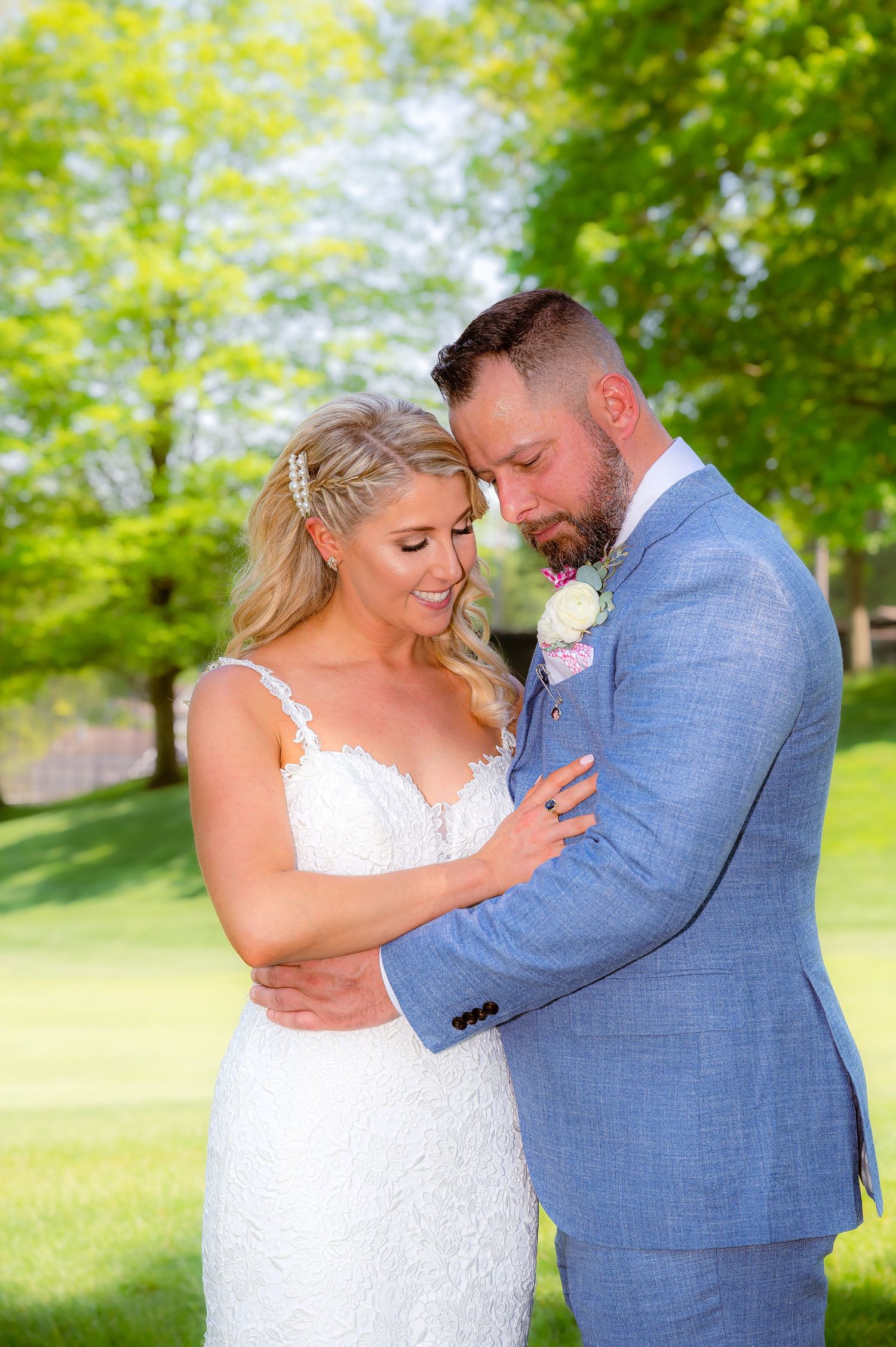 A romantic outdoor wedding photoshoot with a couple in a white dress and light blue suit embracing on a sunny lawn.