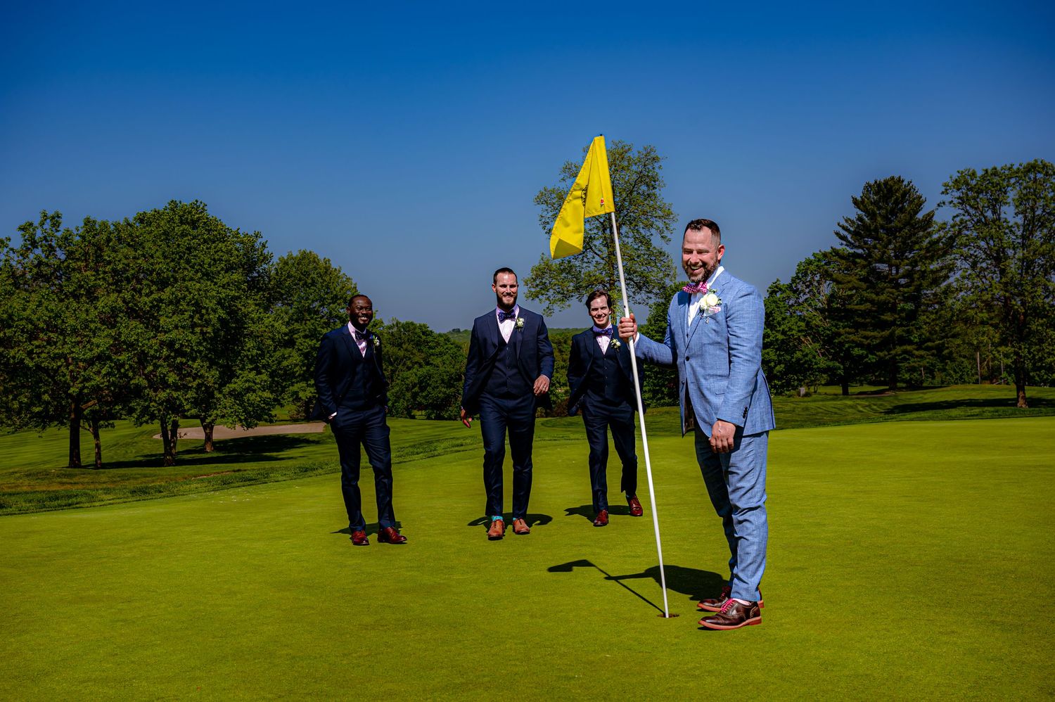Group of people gathered on a sunny golf course with yellow flag pin visible in distance.