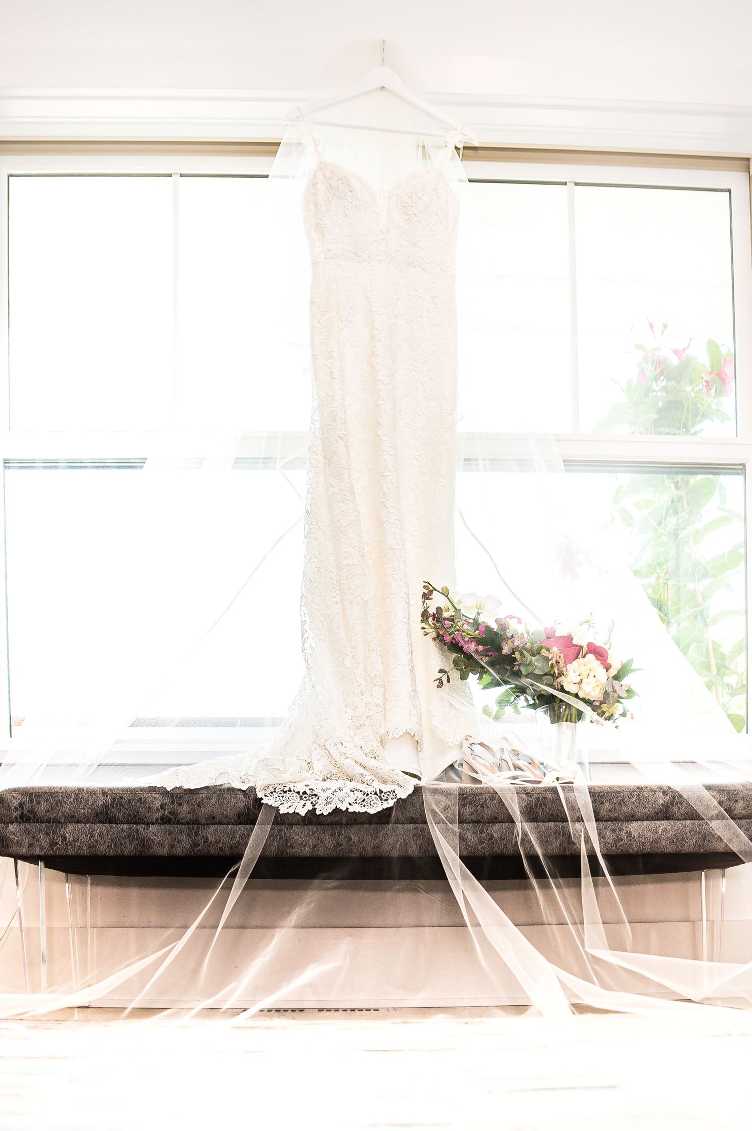 An elegant white lace wedding dress hangs by a bright window with a floral bouquet resting on the windowsill.