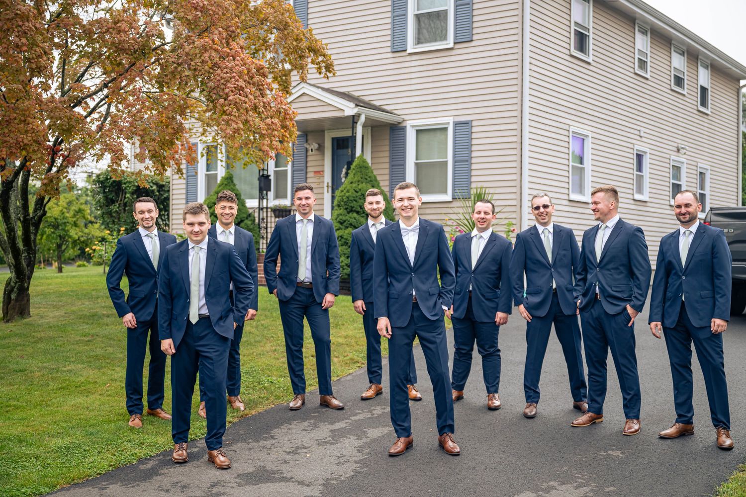 A group wearing matching navy suits and brown shoes poses together on a driveway in front of a gray house.