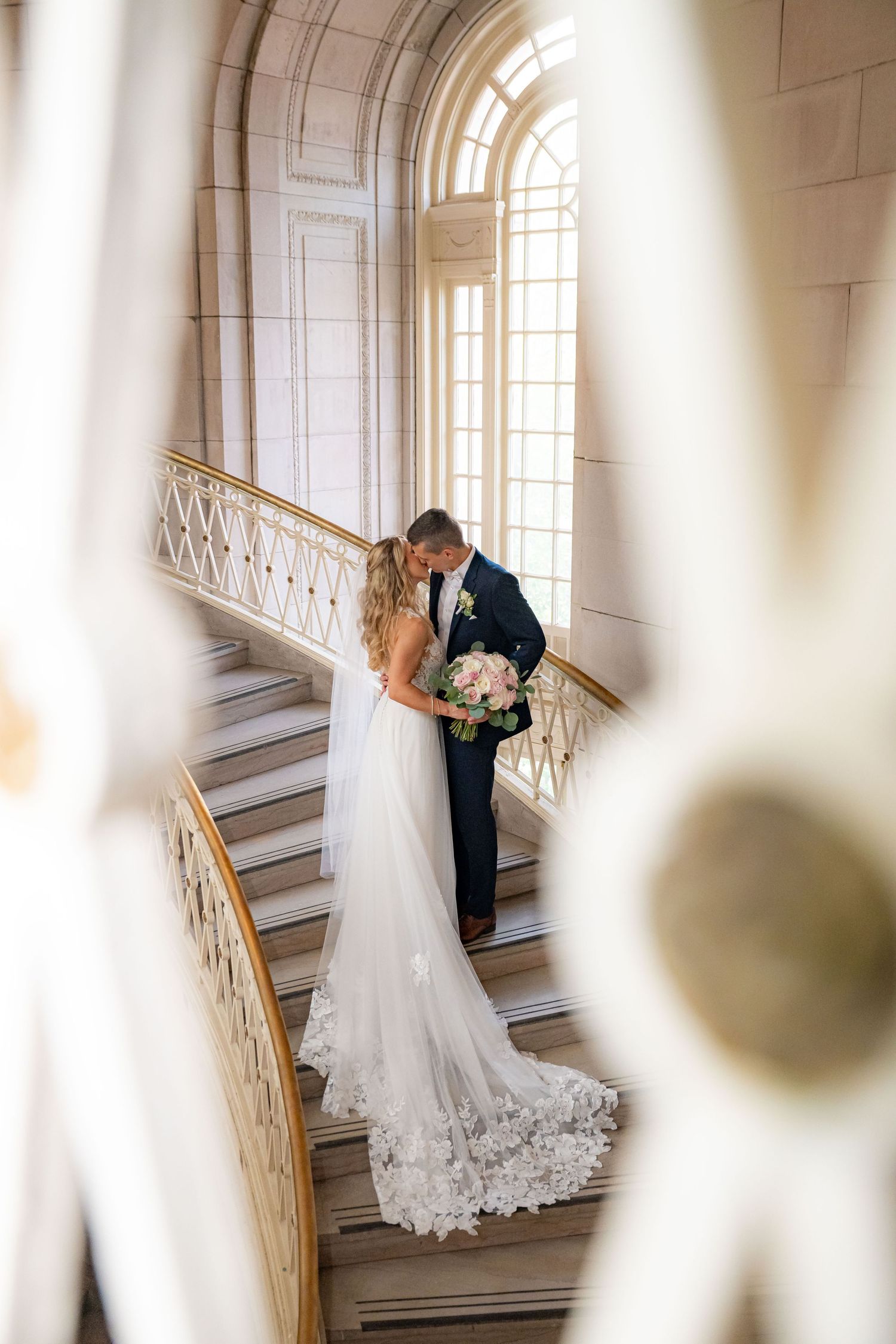 Newlyweds share an intimate moment on an elegant white staircase with arched windows in a historic building.