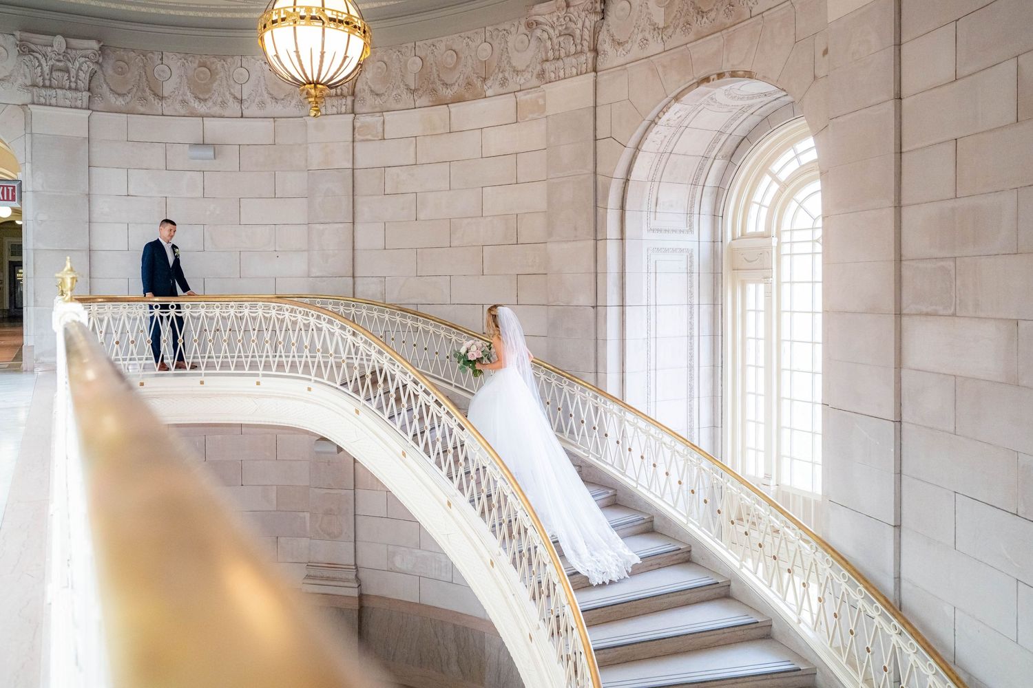 Elegant curved marble staircase with ornate railing in grand hall with arched windows and pendant lighting.