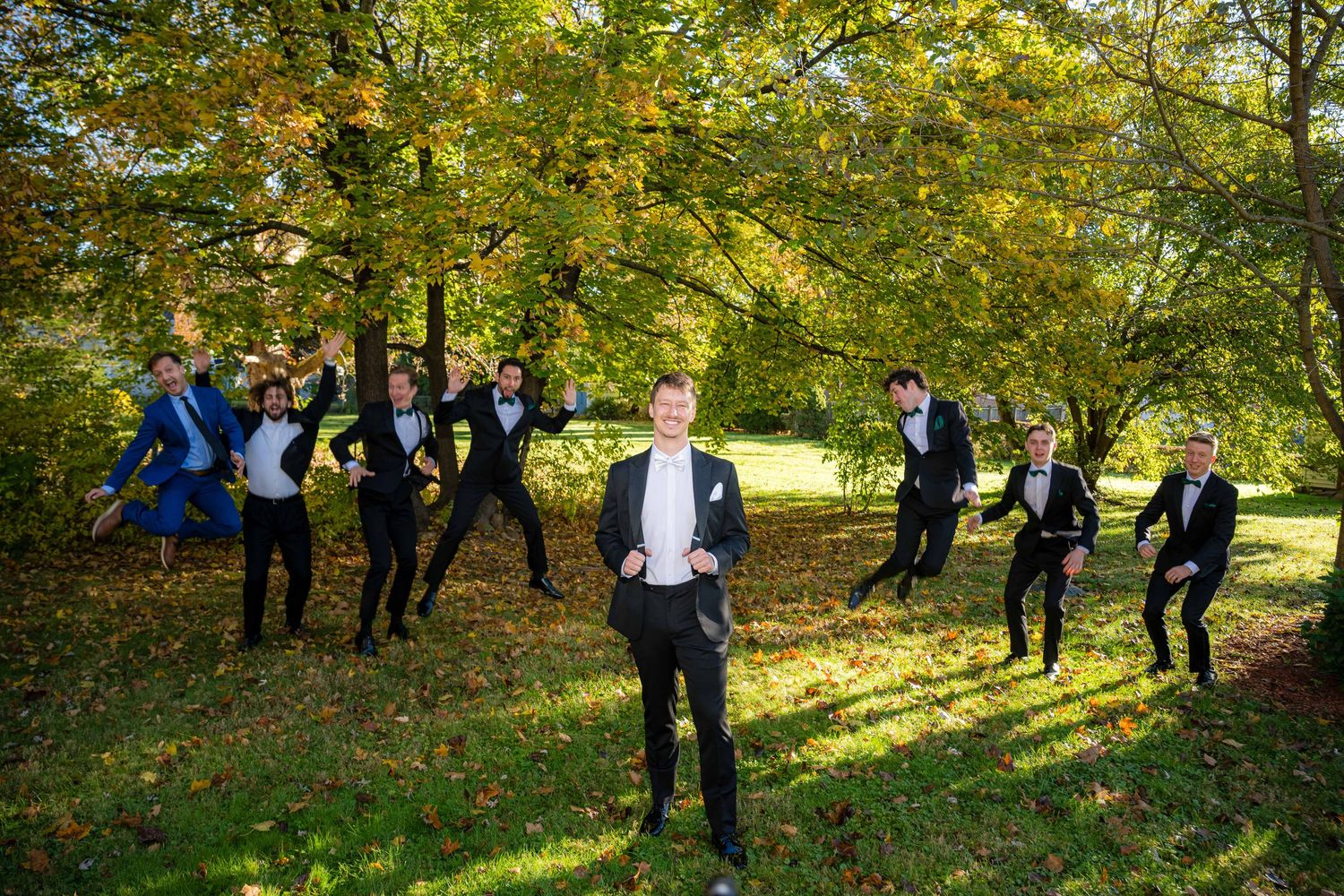 A group of people in formal attire pose for photos in a sunny autumn garden setting with golden foliage.
