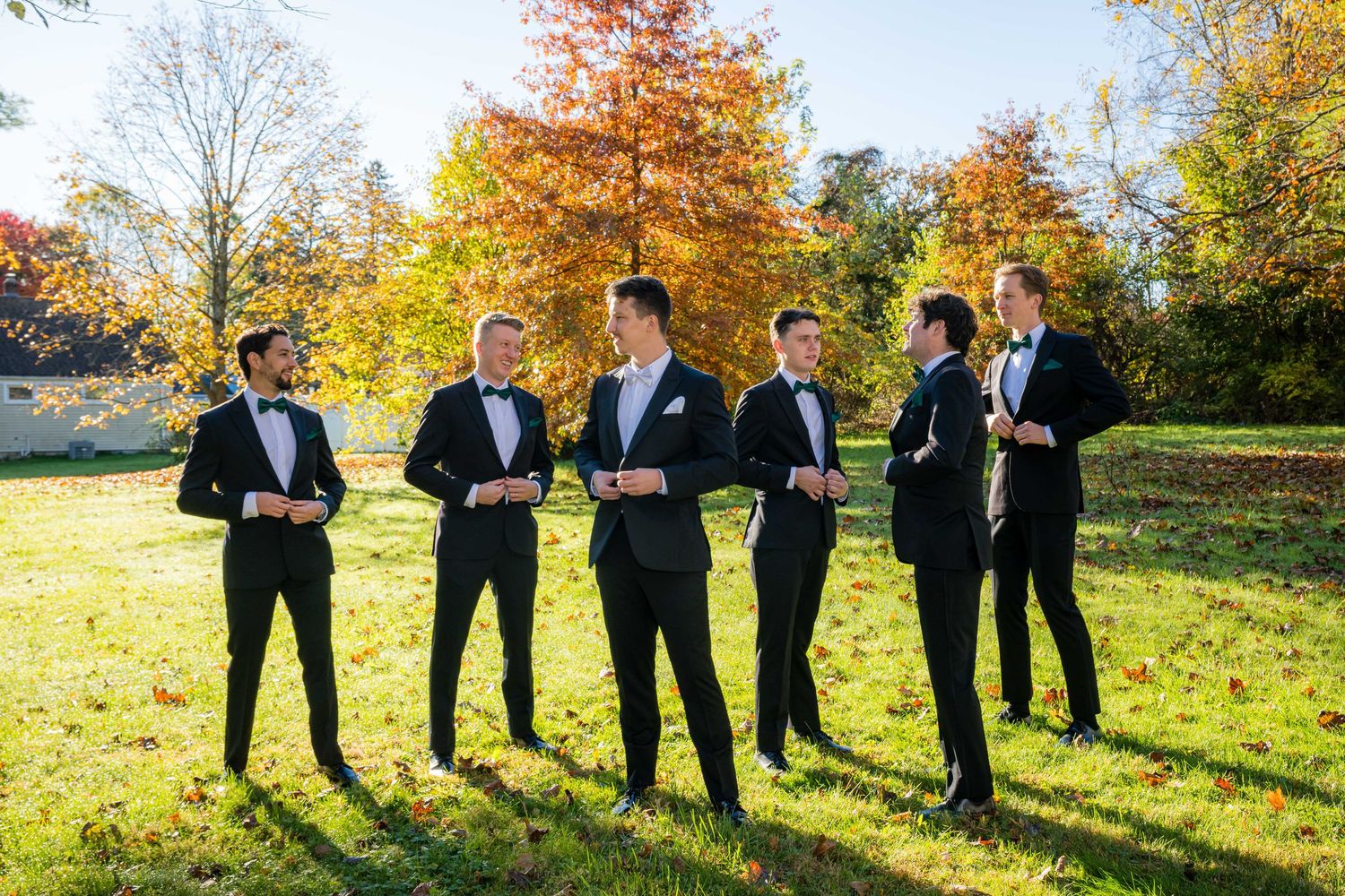 A group in formal black tuxedos stands together on a sunny autumn day with colorful fall foliage in the background.