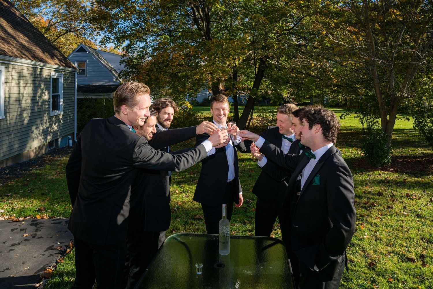 A group in formal black suits celebrates outdoors with champagne bottles on a sunny autumn day.