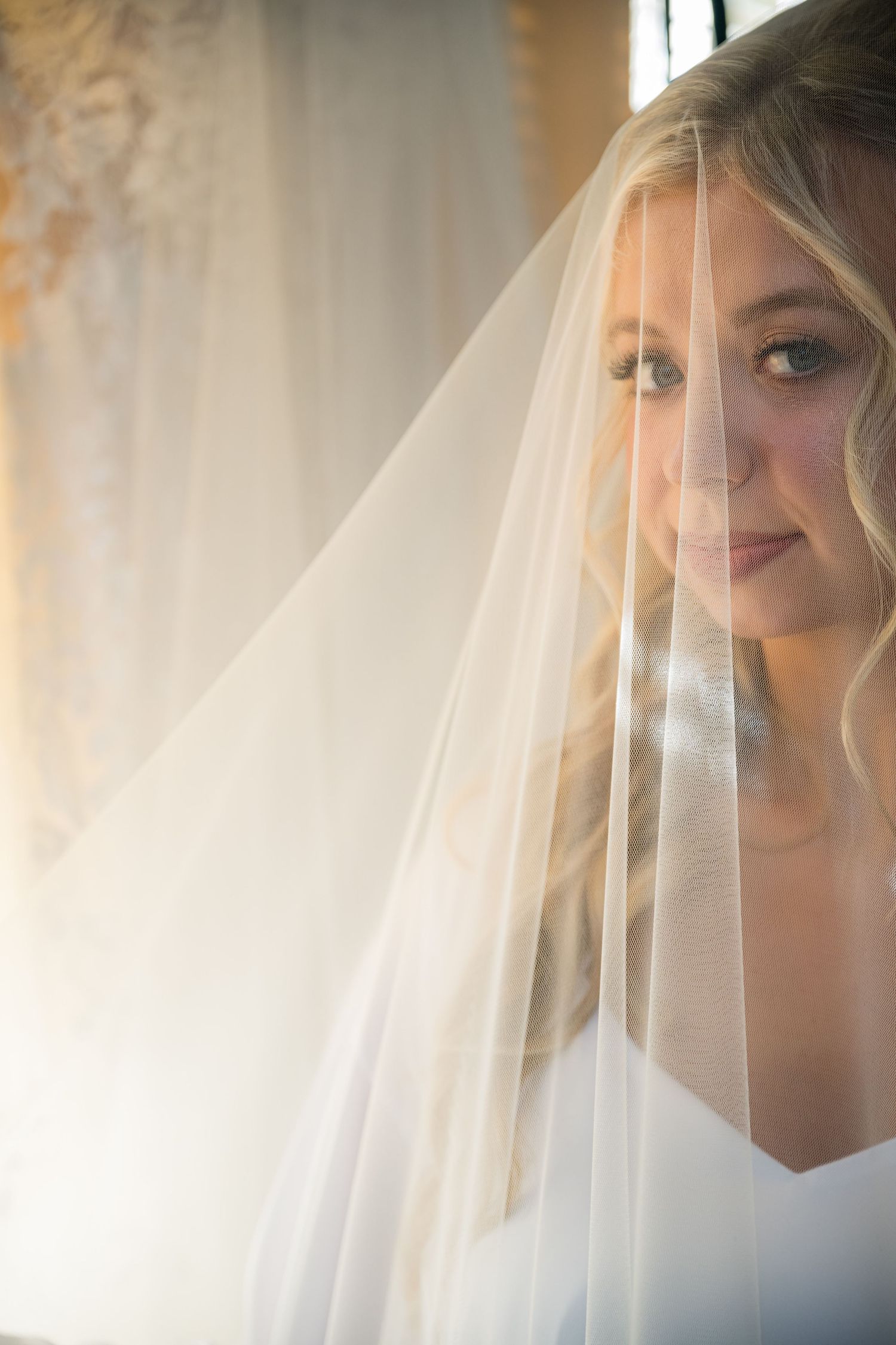 A bride in a delicate white veil smiles softly in warm natural window light during her wedding day.