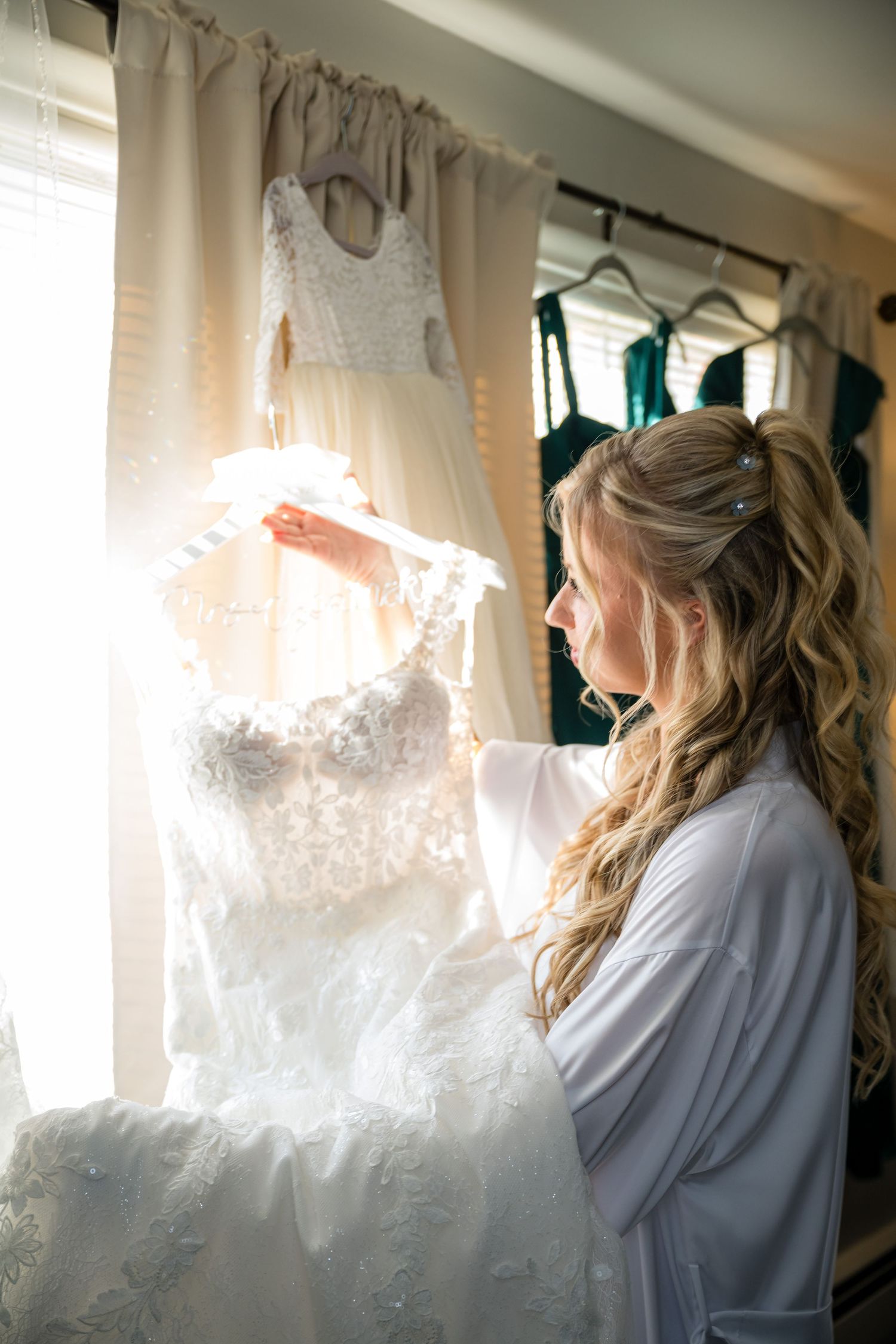 A person in a white robe admires an elegant lace wedding dress hanging by a window in natural light.