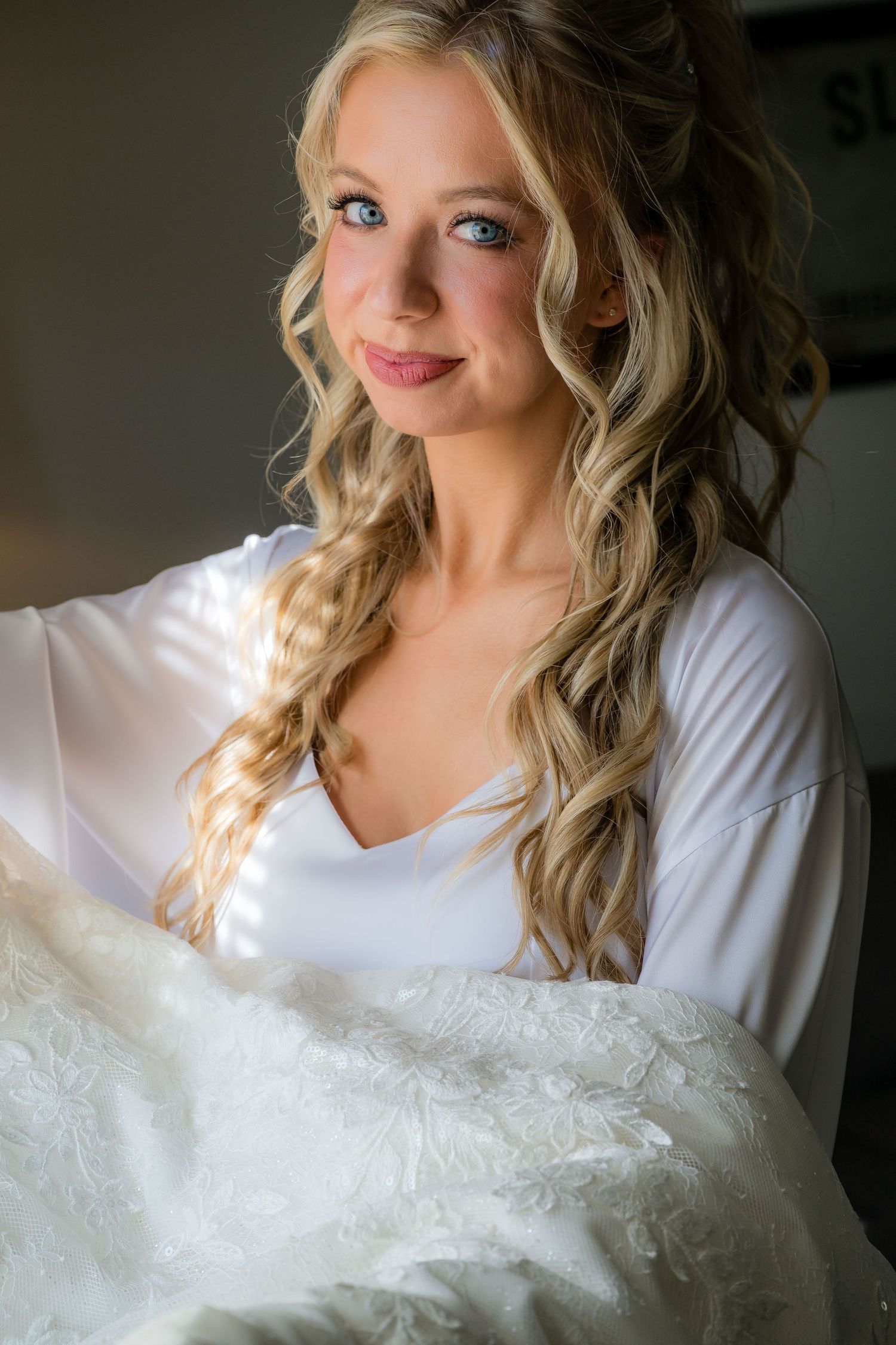 A person with long blonde curled hair wearing a white robe holds a white wedding dress in soft natural lighting.