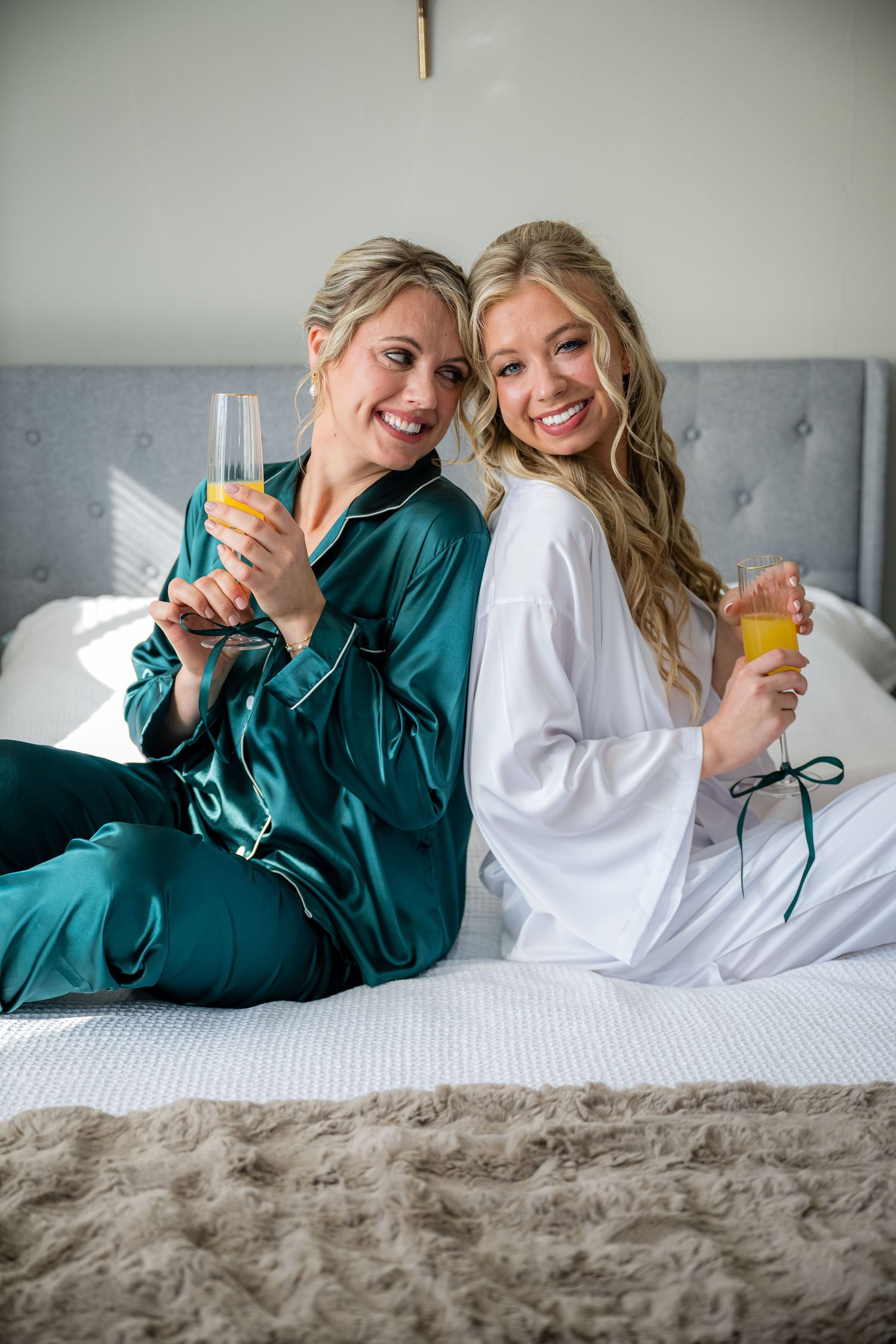Two friends in white and teal loungewear share laughter while relaxing on a gray upholstered bed.