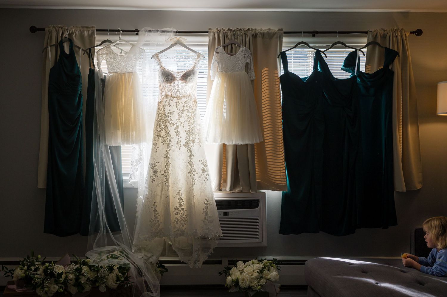 Wedding dresses and formal attire hanging in silhouette against a sunlit window with soft, ethereal lighting.