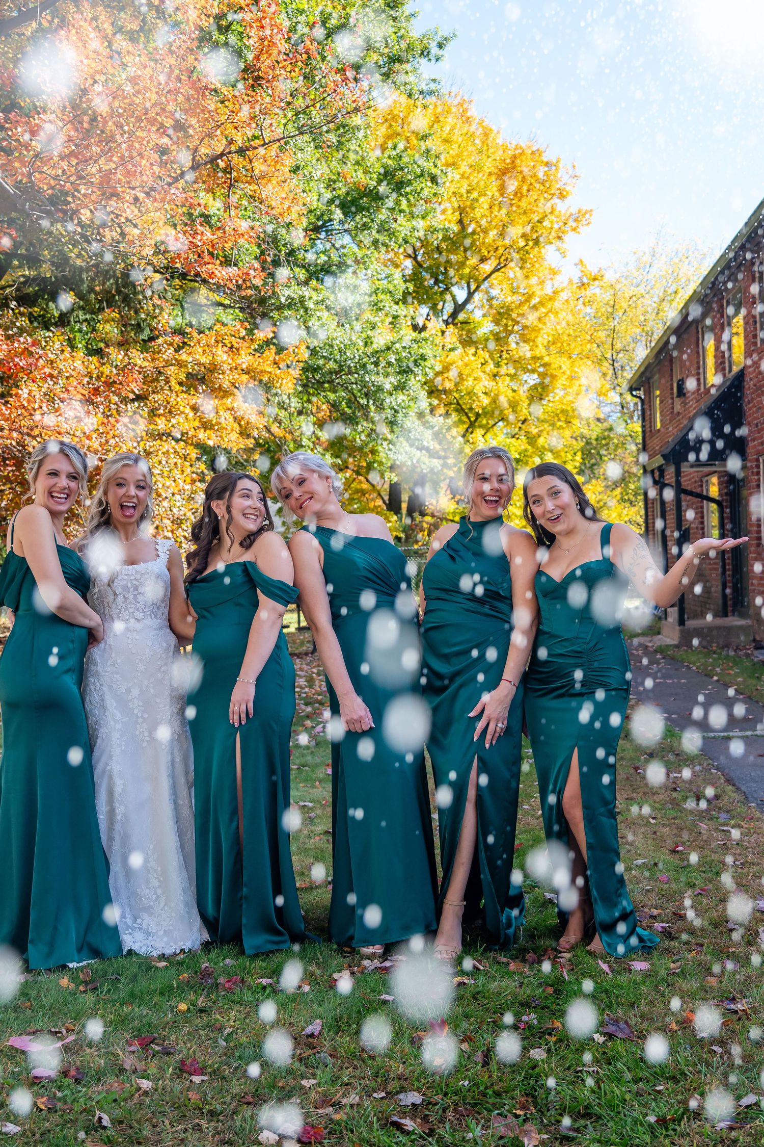 Wedding party in emerald green dresses poses outdoors with autumn foliage and floating bubbles.
