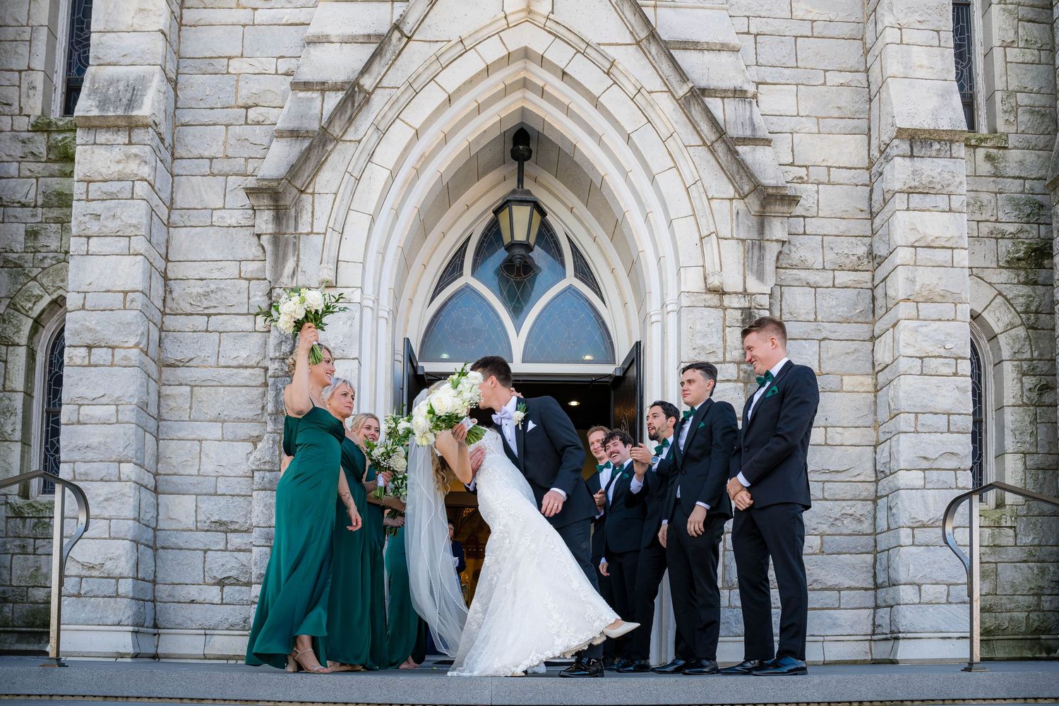Wedding party gathers outside stone church entrance with gothic arched doorway and architectural details.