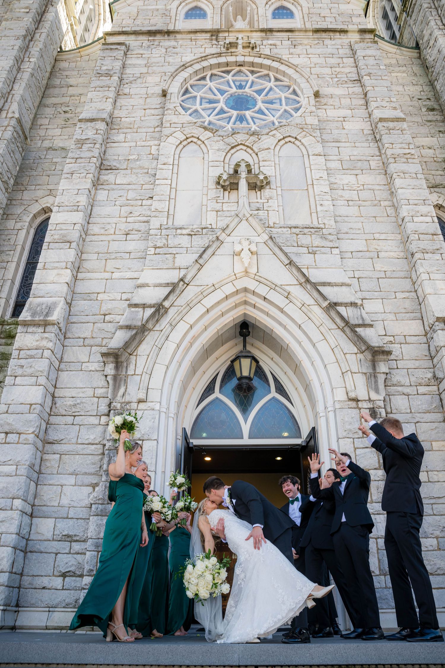 Wedding party poses outside a beautiful gothic stone church with arched doorways and stained glass windows.