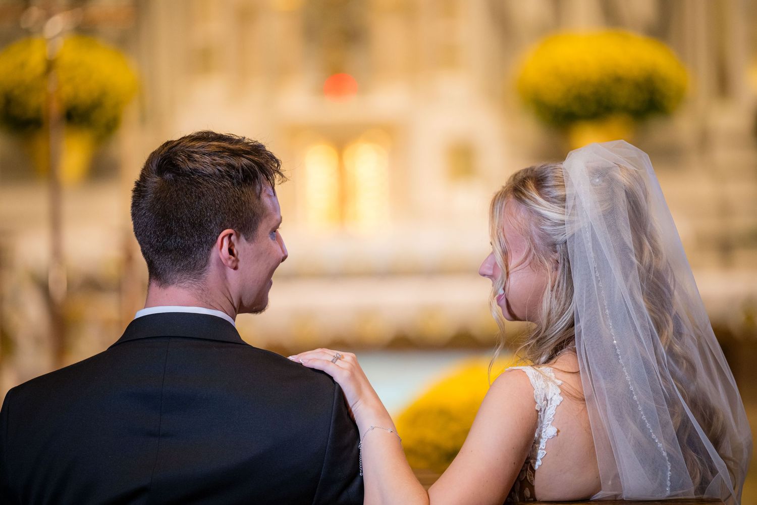 A couple in formal wedding attire gazes at the altar surrounded by yellow floral decorations during their ceremony.