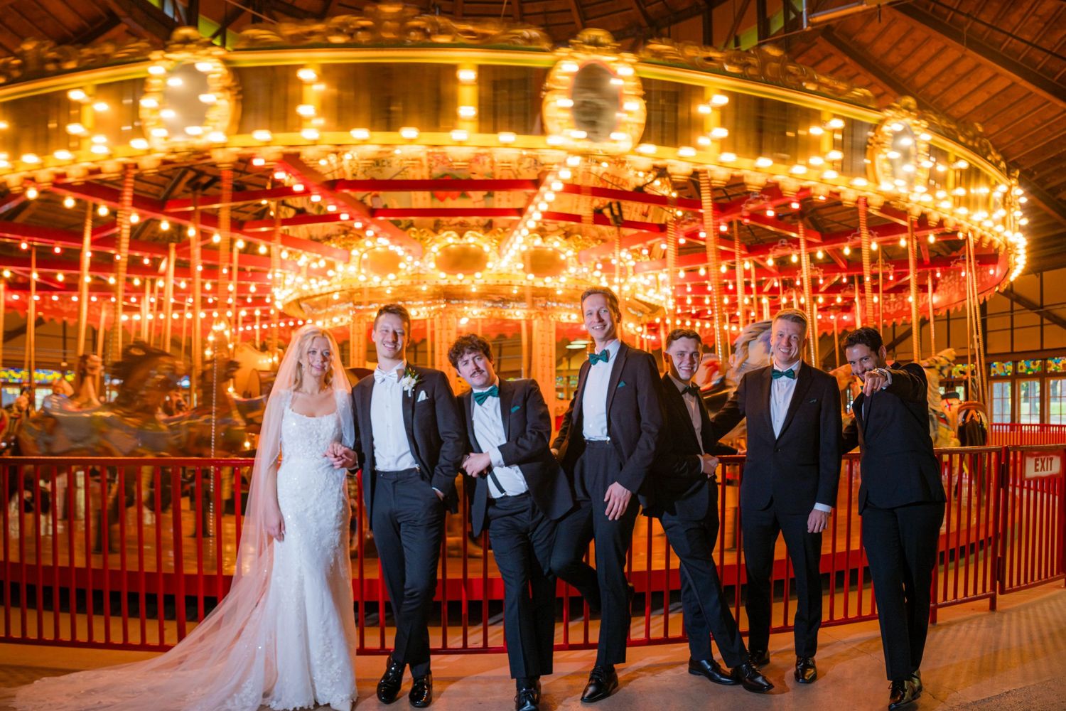 A wedding party poses in front of an illuminated vintage carousel with glowing lights and warm ambiance.