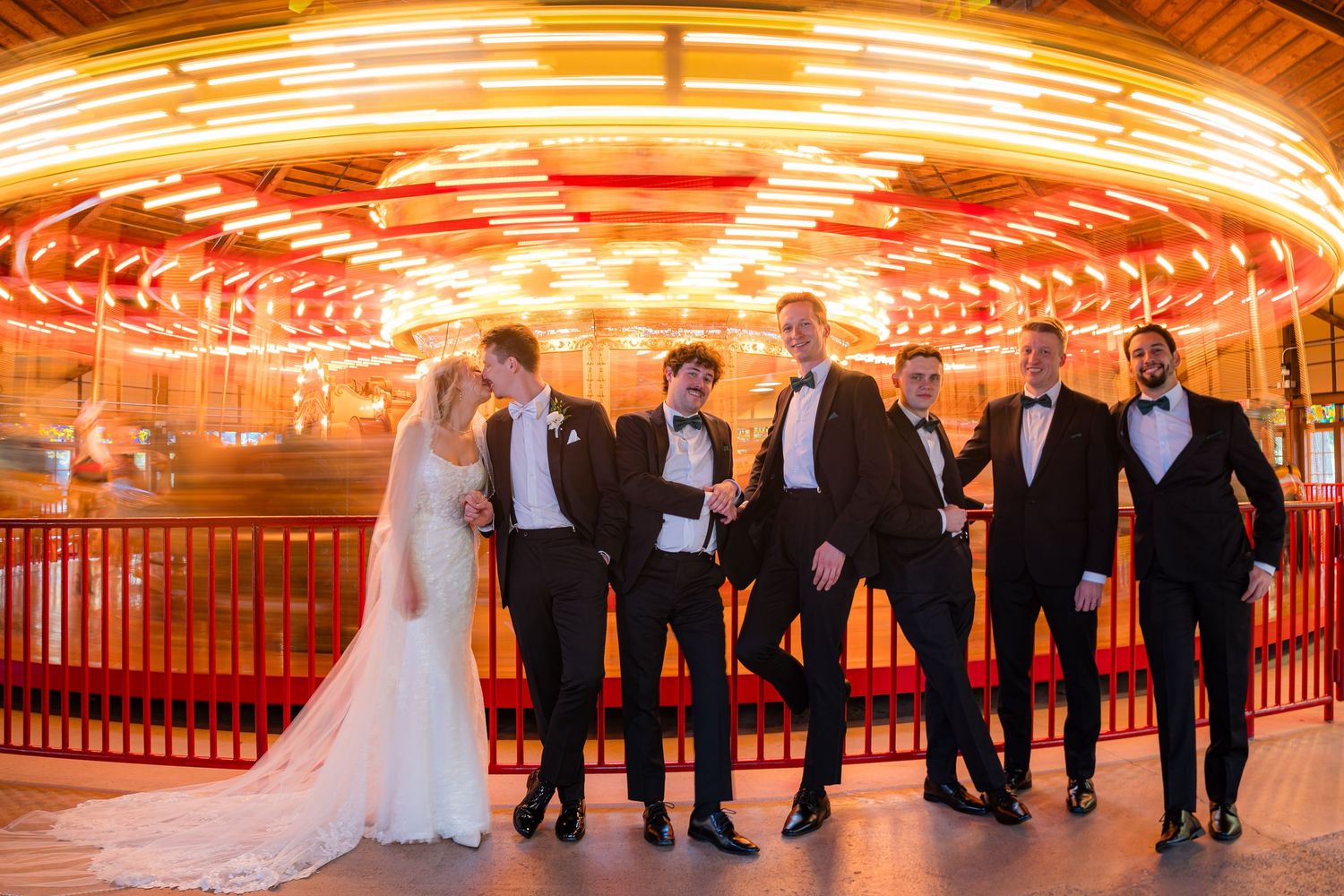 A wedding party walks in front of a glowing red and orange carousel at night creating a dramatic silhouette effect.
