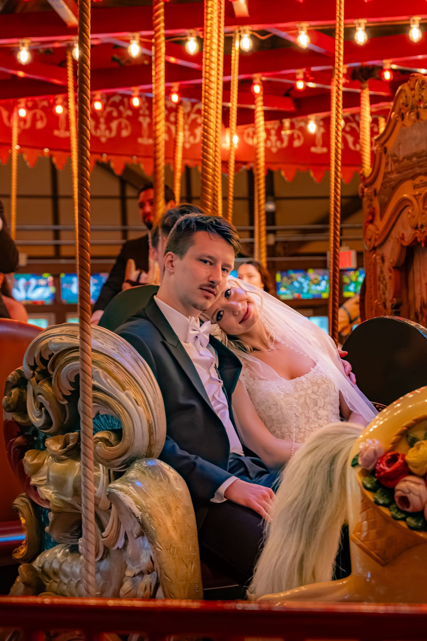 A couple sits together on an ornate carousel with red lighting and string lights creating a romantic atmosphere.